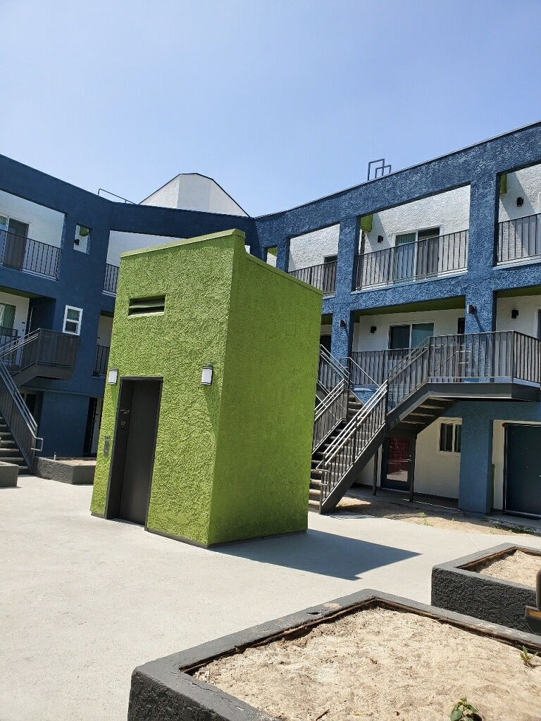 Side view of green elevator. Behind it is a three story white and blue building with a flight of stairs. There are small square areas surrounding the area that are filled with sand.