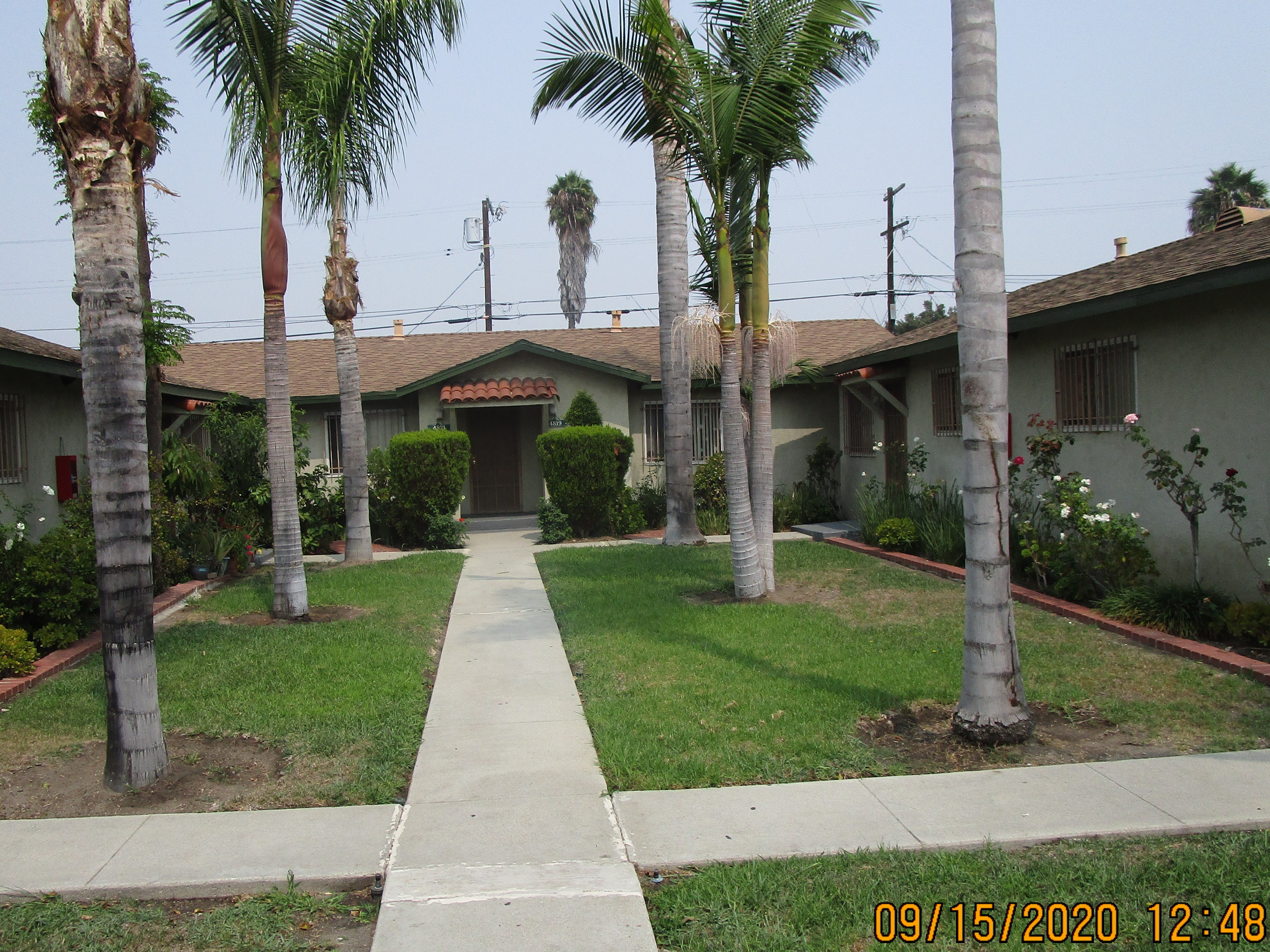 Front view of a courtyard style building, palm trees on each side, roses, plants and bushes around.