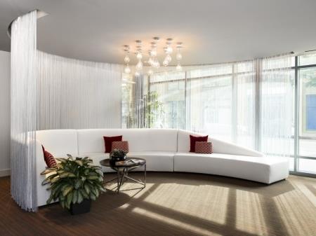 Lounging area with a large curbed sofa and a curtain behind it. Room has large windows, a coffee table and a plant.