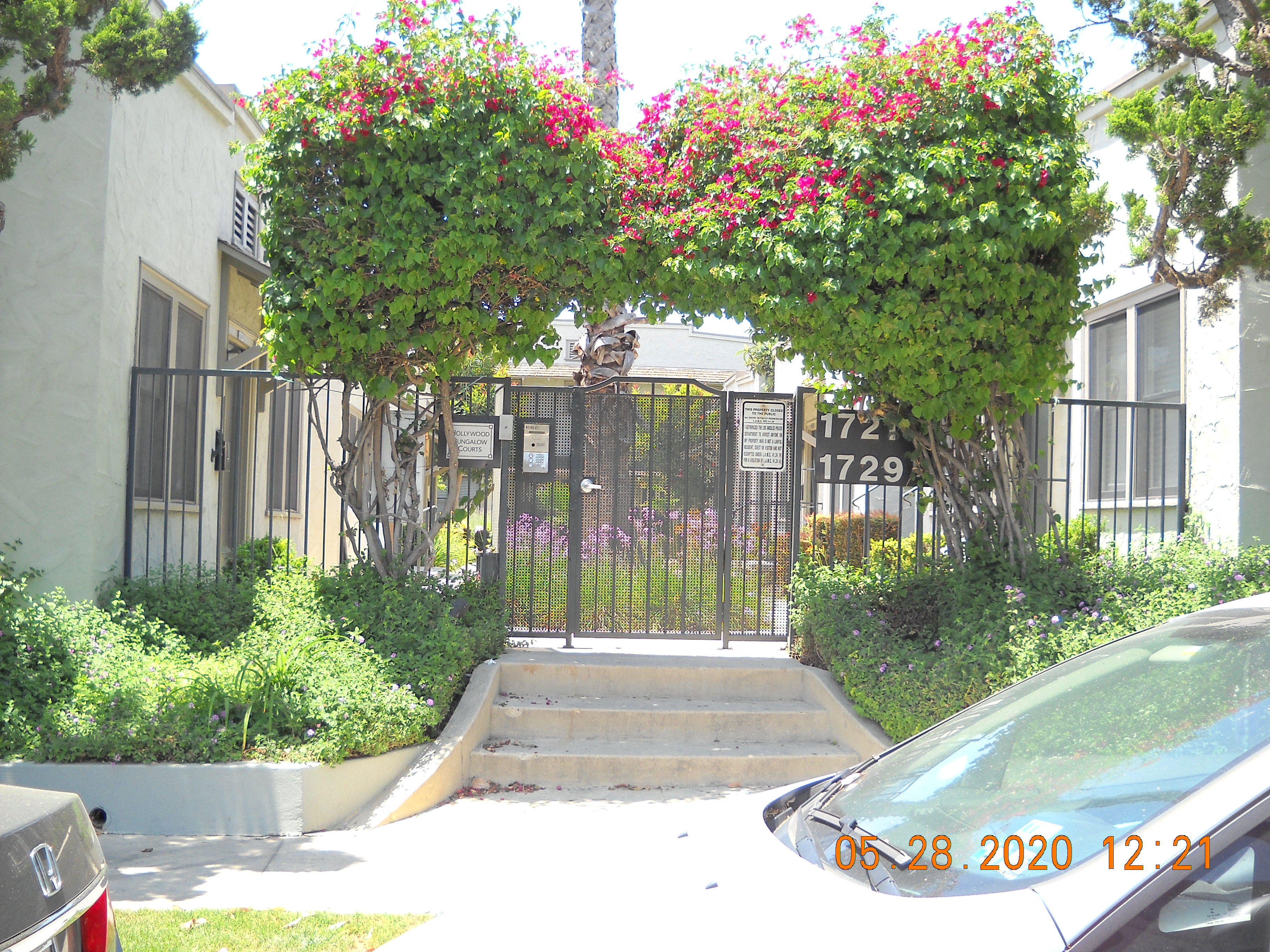 Front view of a gated complex. There is a small staircase leading up to the front gate. There is a keypad. On the sides of the stairs are bushes and trees. Inside the fence are also plants in the center of the passage way leading up to the units.