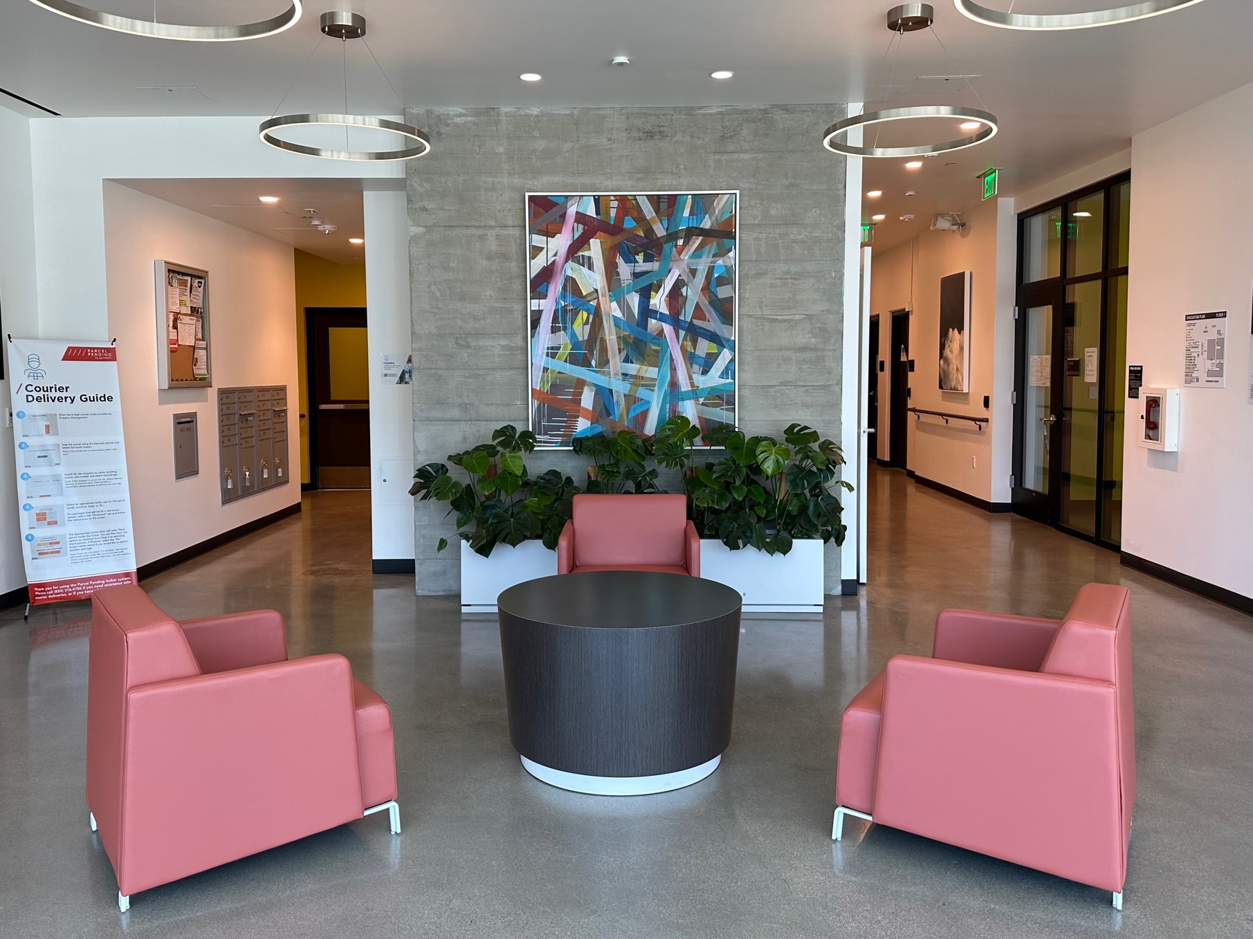 Property Pictures of lobby with circular ceiling fans, three pink chairs surrounding round black table. View of wall with multi colors and a long planter beneath with green plants.