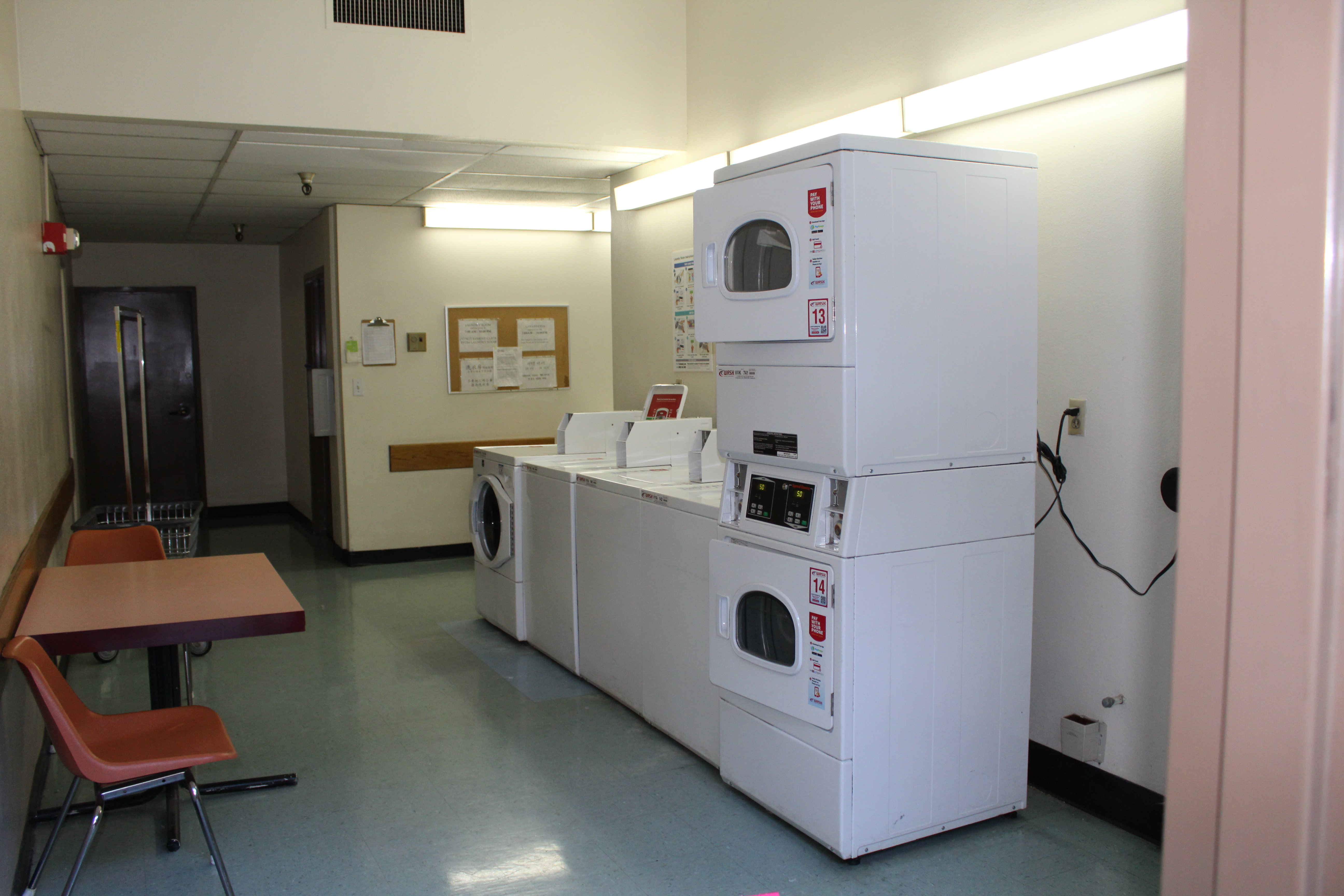 Interior view of a laundry room for Angelus Plaza 1. Three white washers and three white dryers, a square table with two chairs in a rectangular room.