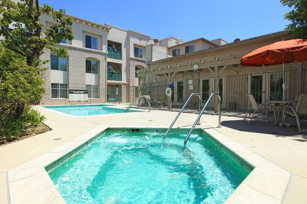 Exterior view of Valley Village Senior Apartments. Jacuzzi and pool area with umbrella table and chairs. 3 story building behind pool area