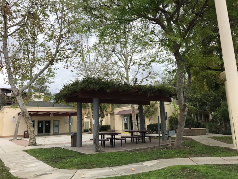 Common outdoor area with 2 covered outdoor bench tables, trash cans, trees, and grass areas. One sory building behind courtyard.