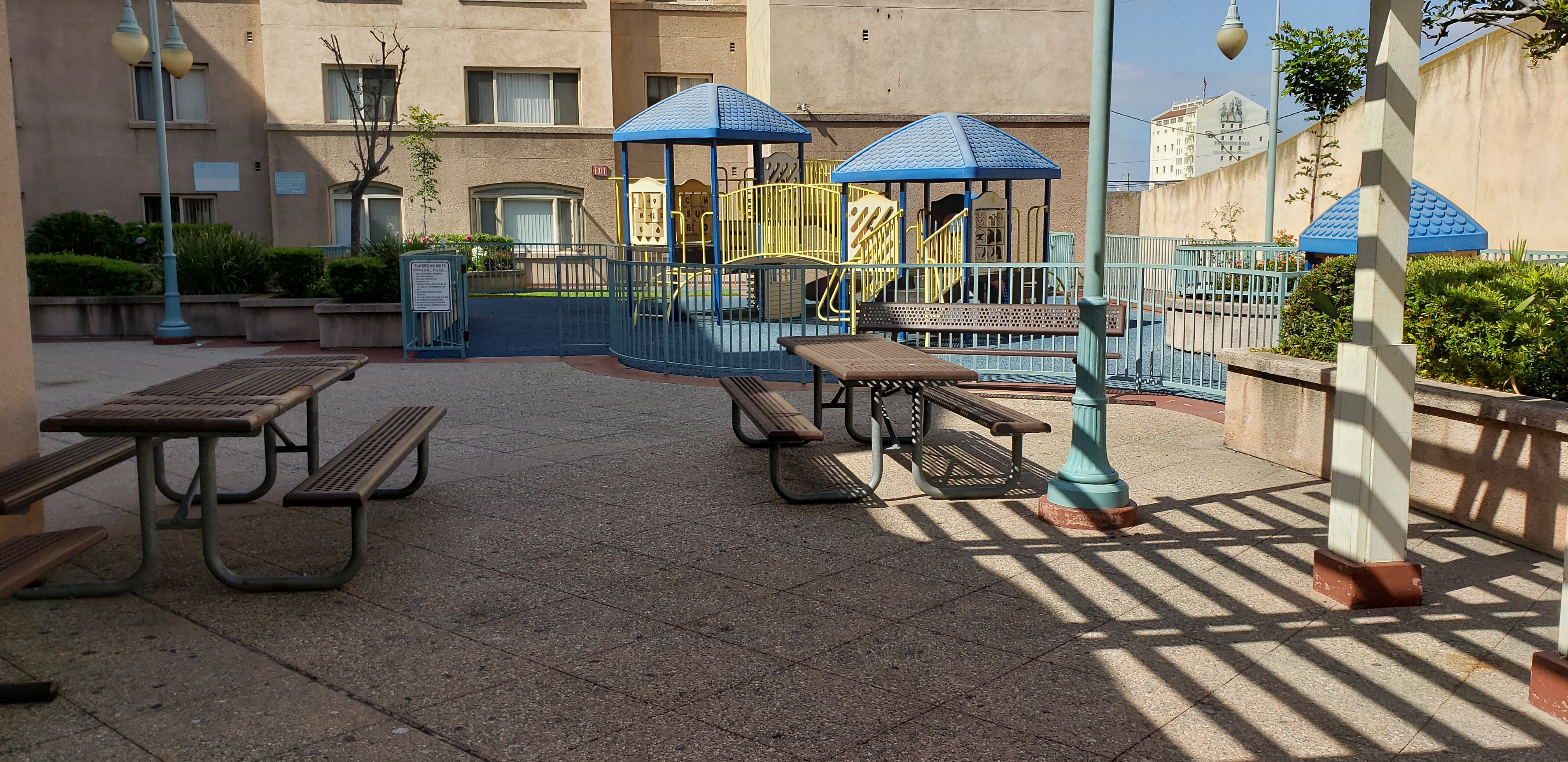 View of a fenced playground with padded flooring. There are a couple bench tables and a bench in front of it. There are plants and bushes around the area.
