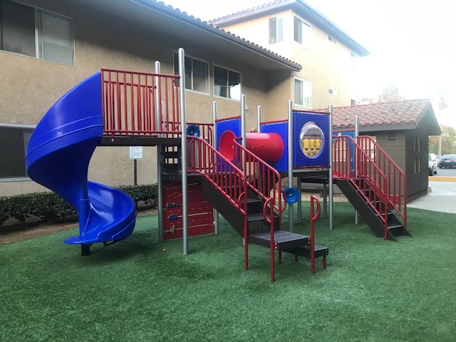 Exterior view of New Harbor Vista Playground. Blue slide on one side with tic-tax-toe game on the other, with black stairs and red hand railings. The playground is on grass and the building behind