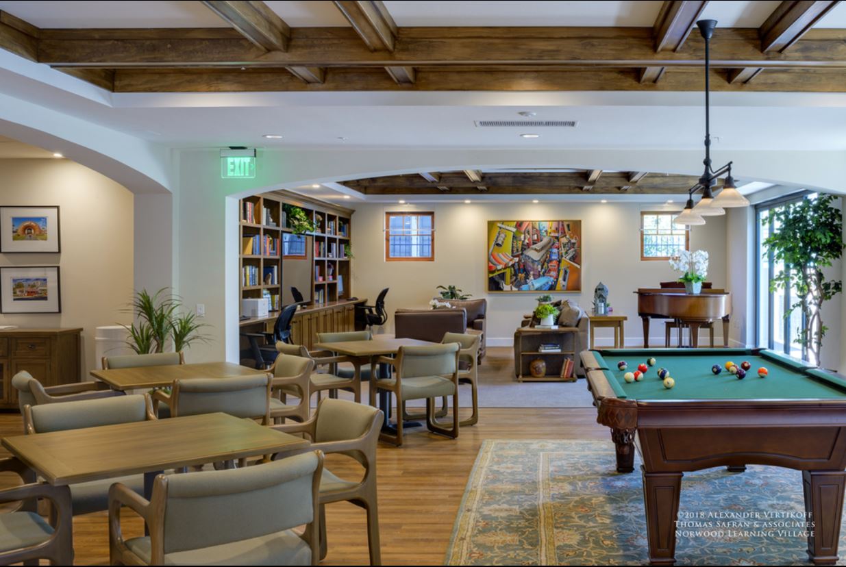 Inside view of a community room. There is a pool table, three table sets, a piano, a computer setup desk with a printer, a bookshelf full of books, and sofas. Across the room there are some plants and modern art on the walls.