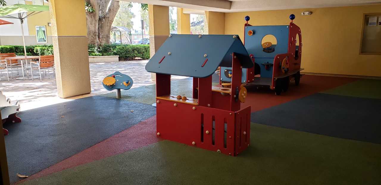 Rayen Apartments playground area. Childrens play area located in a pocket area of the building providing shade. Play area has softpave flooring in multiple colors. Small stationary toy structures. Bulletin board loacted on wall next to play area