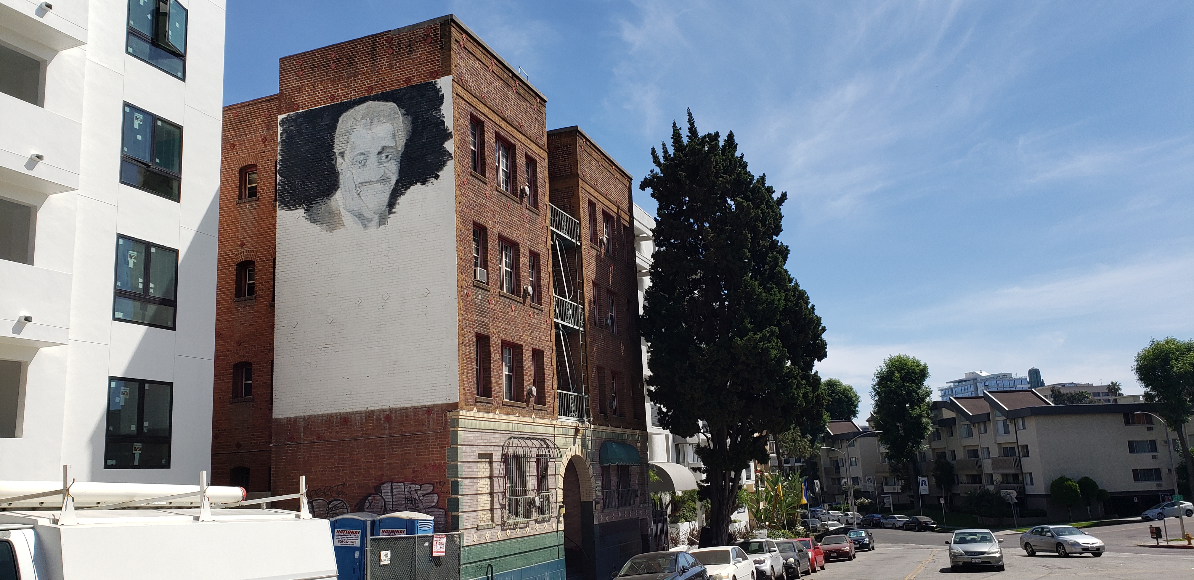Street view of Reno Apartments with an unfinished mural of Sammy Davis Jr on the side of the building and large tree in front.