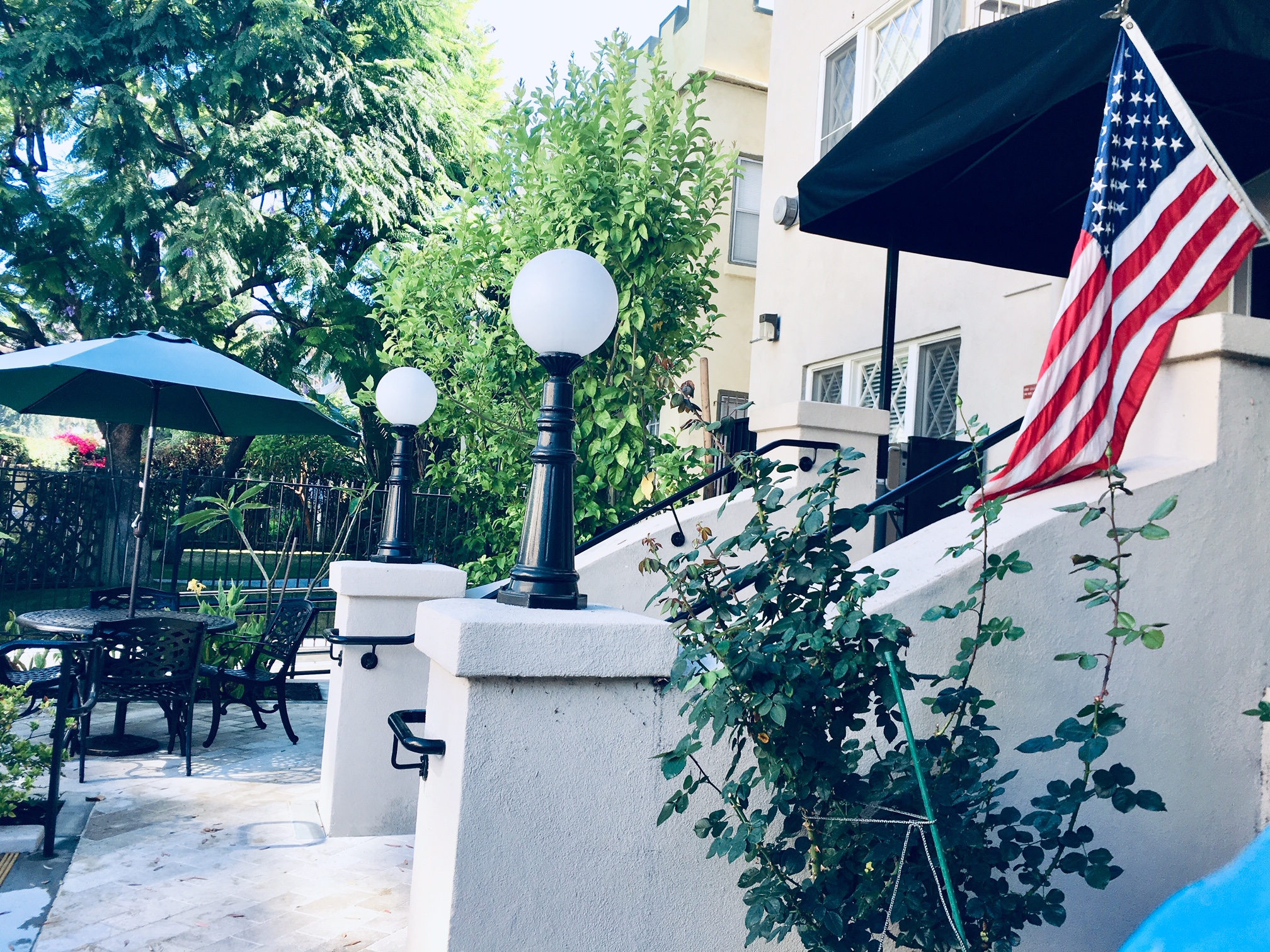 Side view of a patio area. There is a table with an umbrella and chairs. There is an area with newly planted trees. There is a small staircase with handle bars on both side, two lamps, and a small tent canopy with an United States flag on top.