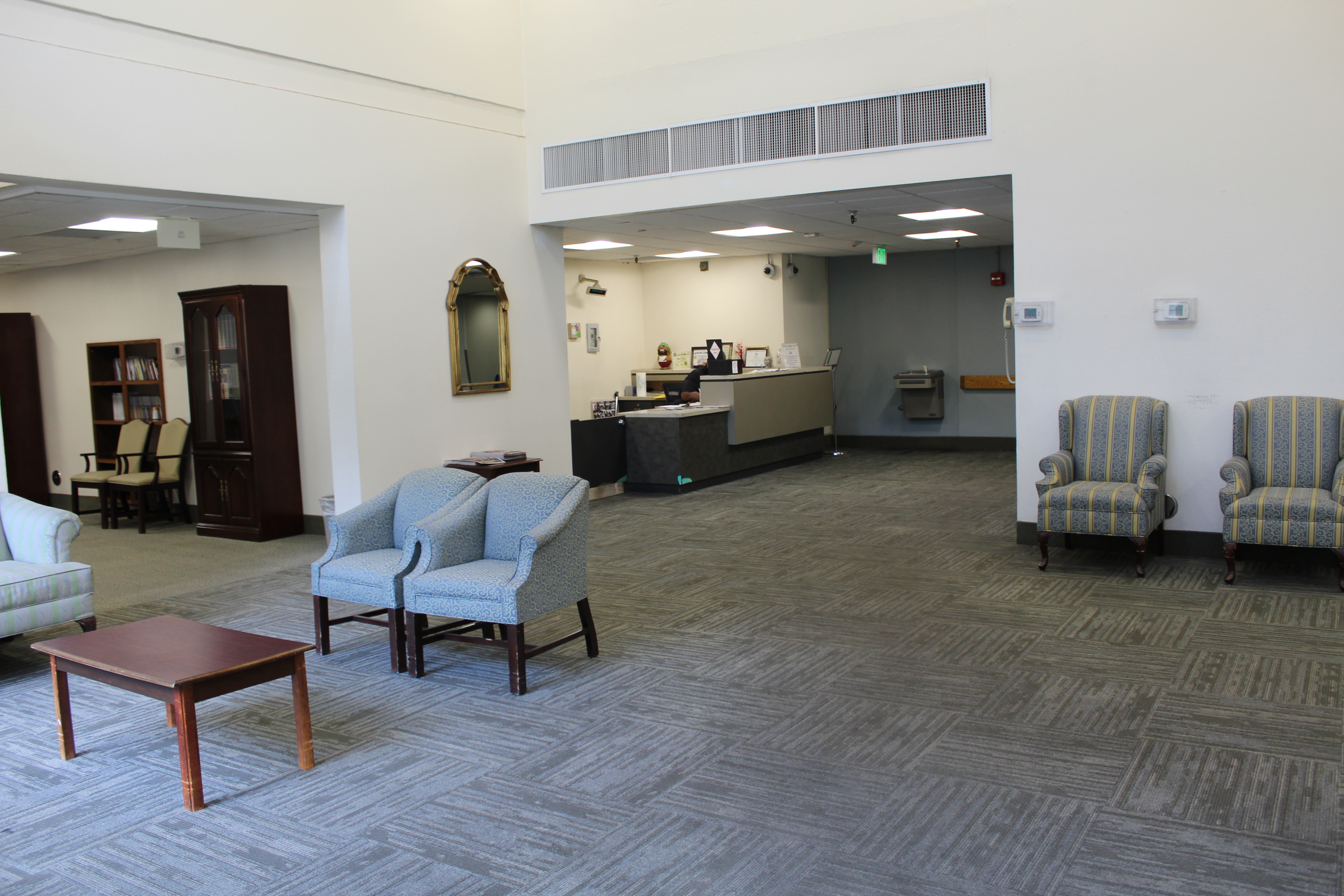 Interior view of community room and Information desk at Angelus Plaza 1. Nuetral colored sofa chairs, brown coffee table, dark brown curio cabinet , and a mirror hanging on a wall