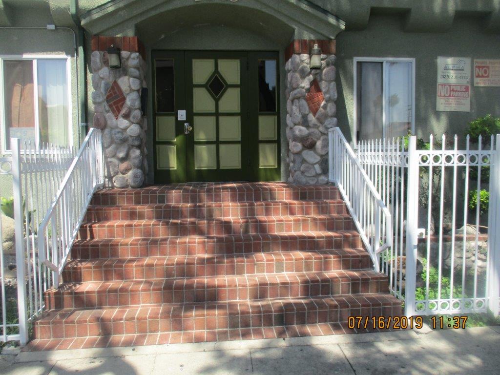Zoomed in main entrance, dark and light green door with triangle and square design, stone pillars on each side of the main door with a lamp on each one, seven brick stairs with white handrails, window on each side of the main door, no public parking sign.