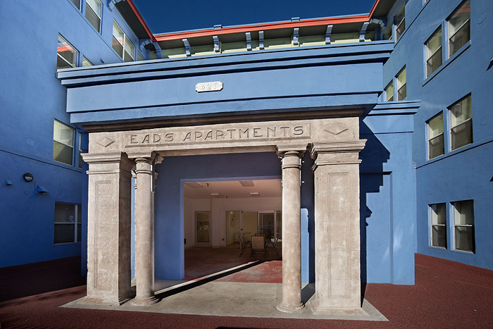 Front view of an entrance to a four story building, Most of the building is blue, middle section is lime green. Entraance is round level. and has pillars and an overhead before entering the lobby area.