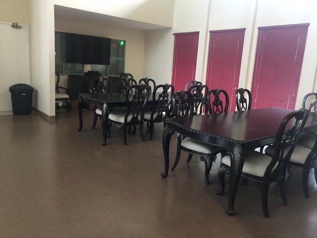 Side view of a dining room, two big dark brown square tables with six chairs each, a white door, a big black trash can with a plastic bag on the left side, three burgundy doors on the right side.