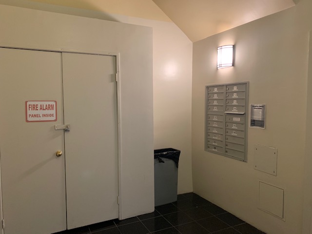 View of a mail room, gray box of 21 mailboxes on the right side, a ceiling lamp on top of the mailboxes, a gray tall trashcan with a black bag, a locked door with a "Fire alarm panel inside" sign on the left side, beige walls.