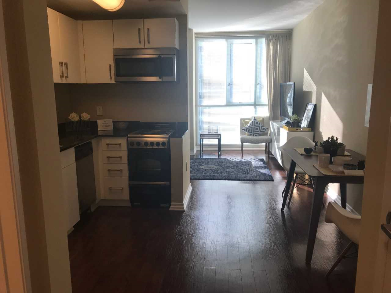 Inside view of a unit from the kitchen area. Kitchen has a small stove, a microwave overhead, and a dishwasher on the side. There is also wood flooring in the unit and has large windows.