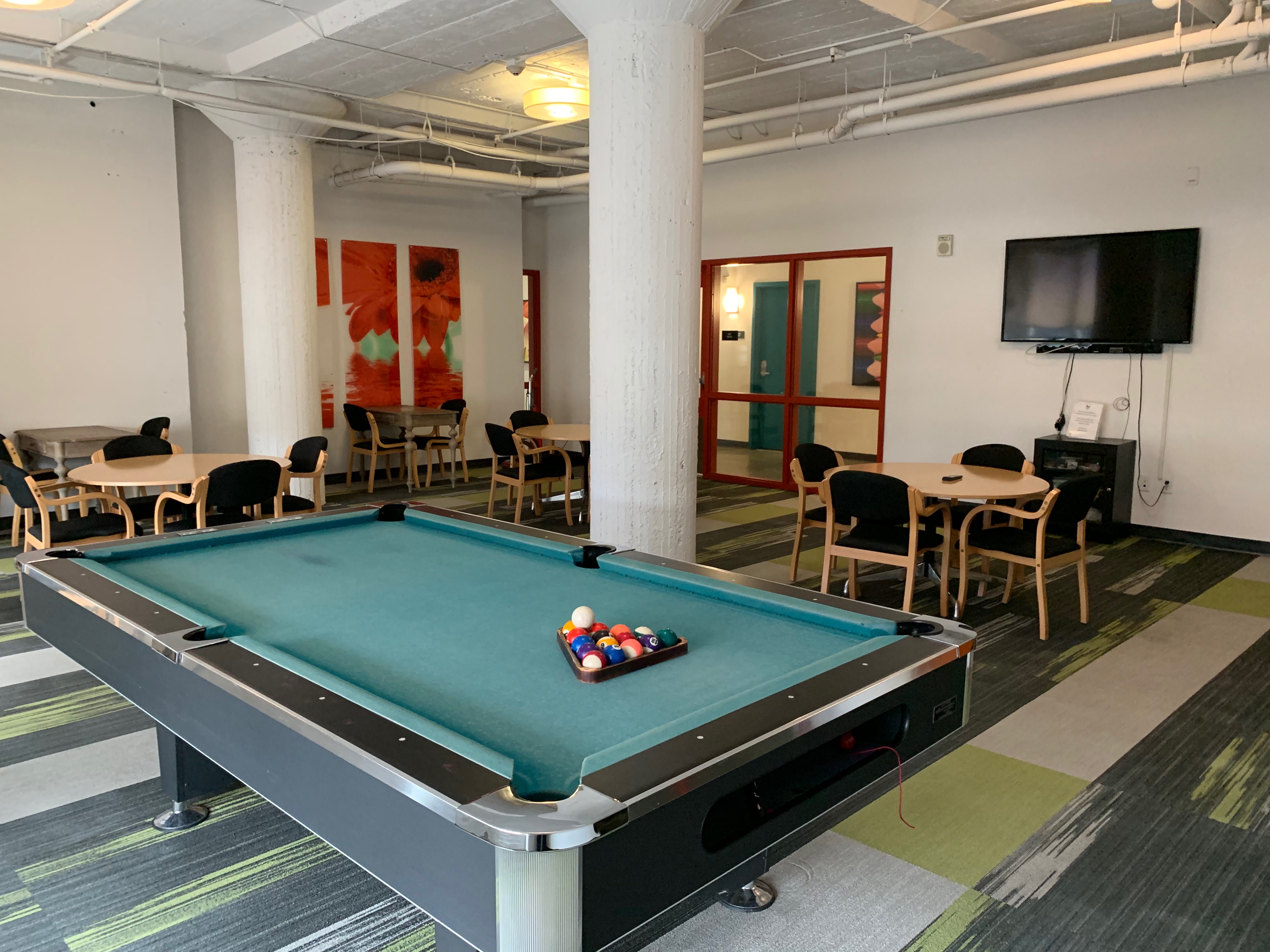 Inside view of a community room. There is a pool table, a flat screen tv, and multiple tables with chairs.