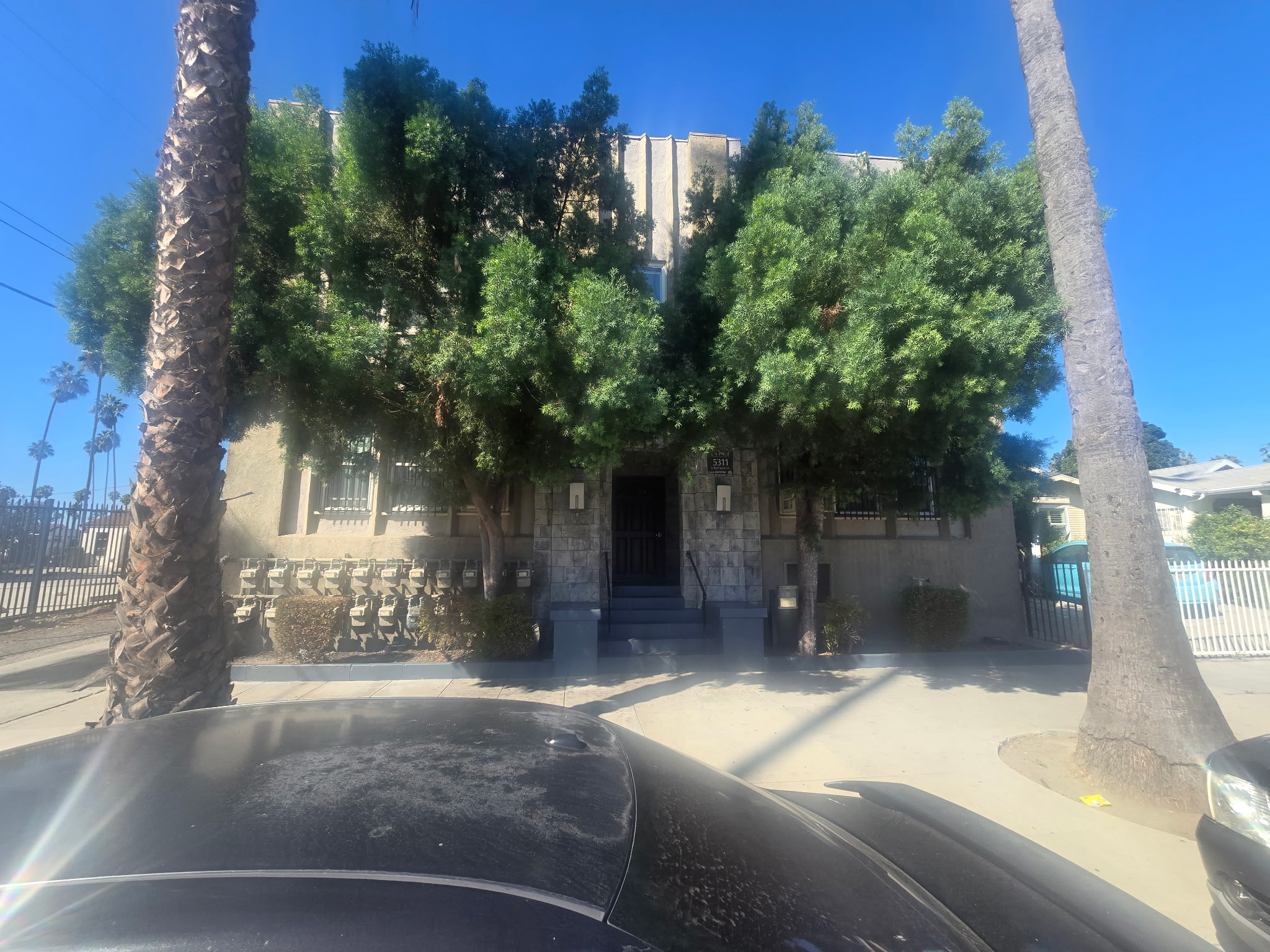 Image of front of building with front door entrance, large palm trees in front of building and vehicles parked on street. 