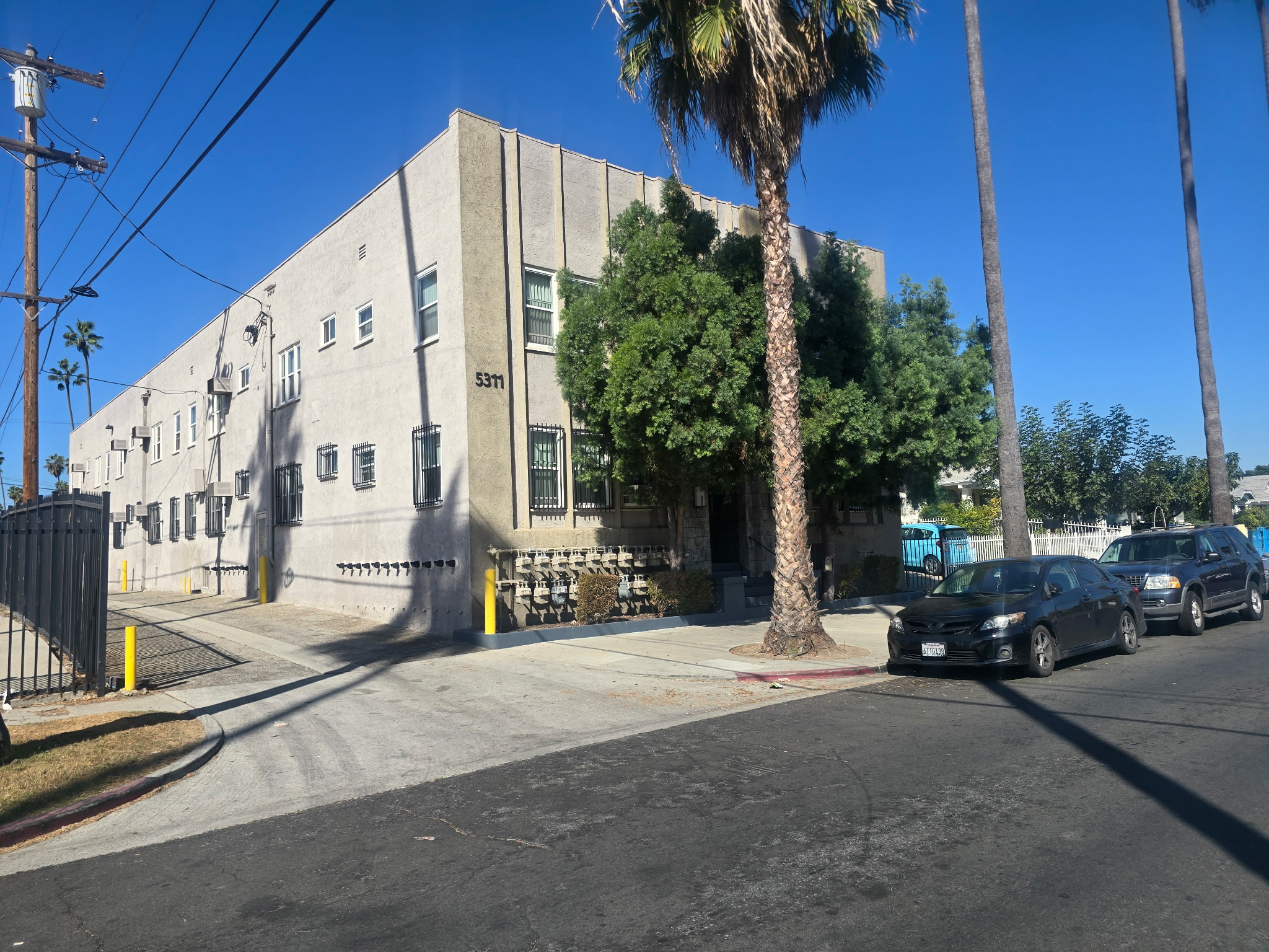View of front of building with adjacent alleyway, large palm trees in front of building and vehicles parked on street. 