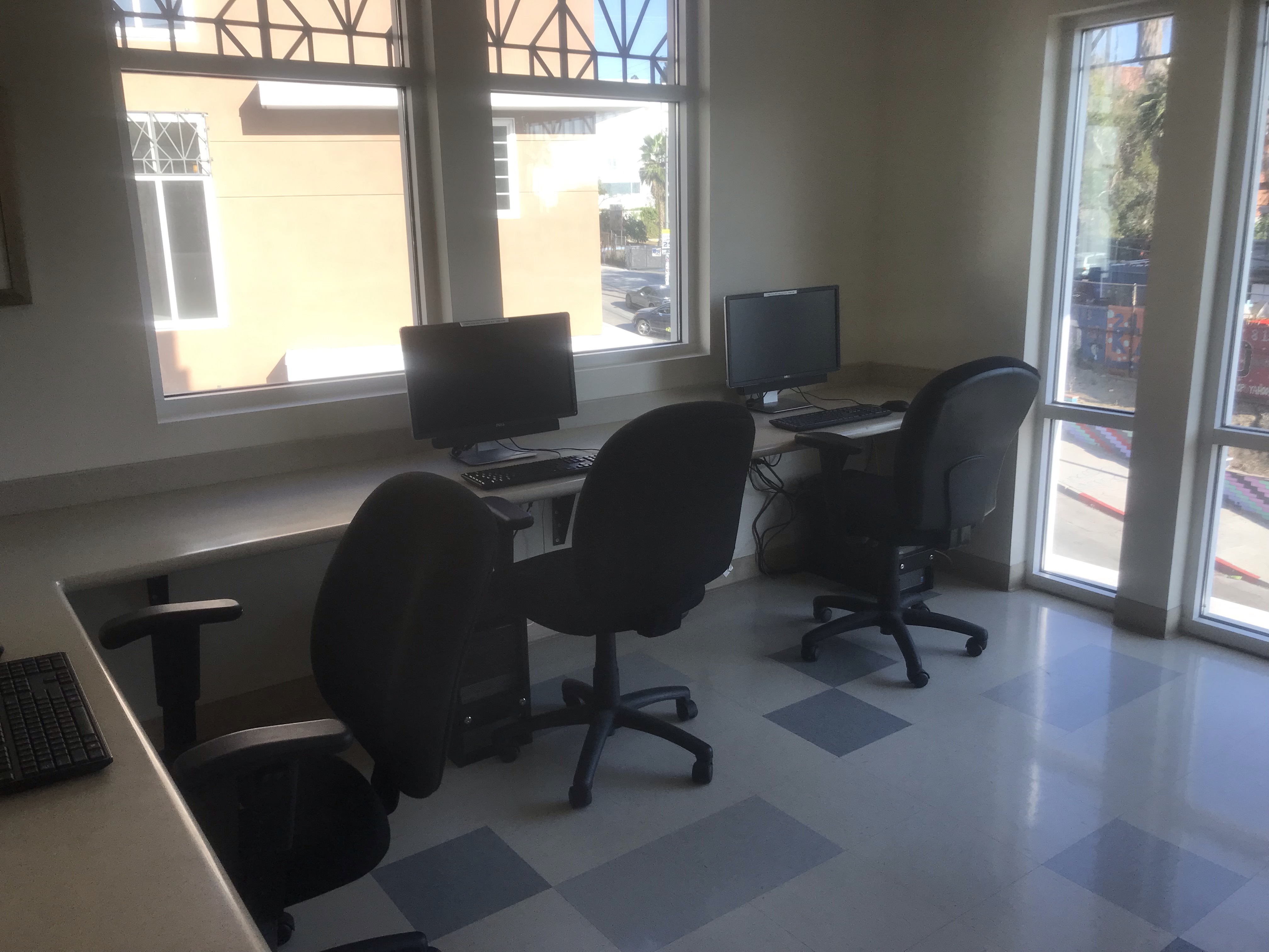 View of a office workstation, three computers, three desk chairs, gray tile flooring, two windows.