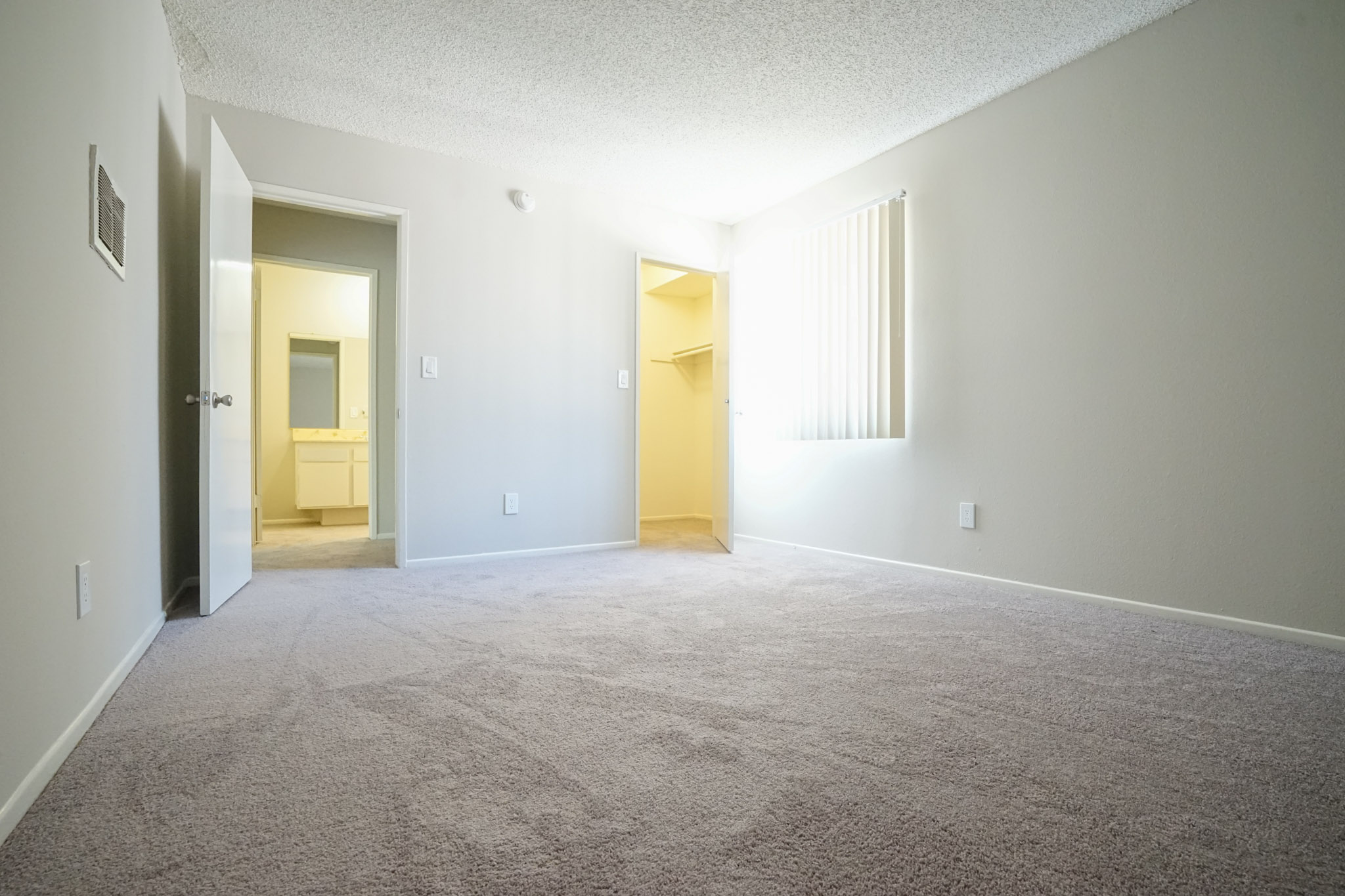Different view of a bedroom with carpeted floors. Room has a window with blinds and a closet with a door. The bathroom is located across the entrance of the bedroom.