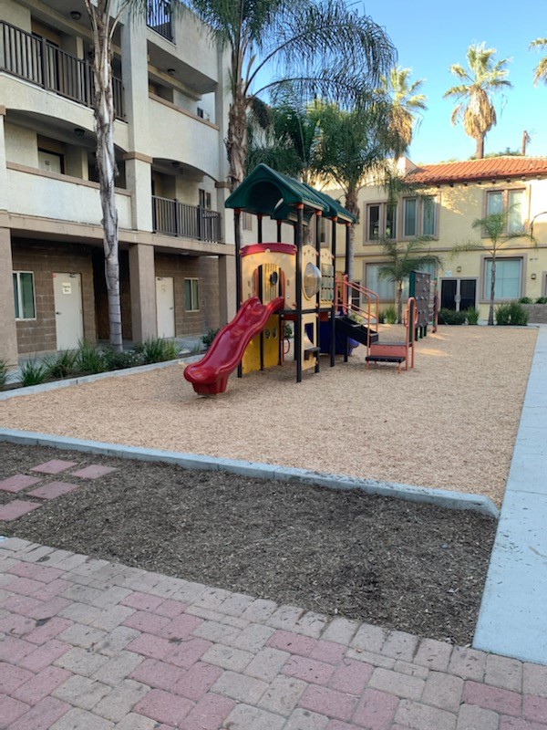 View of an outdoor playground. Visible is a slide, climbing stairs, and a viewing window. There are trees and small plants on the outside perimeter of the playground.