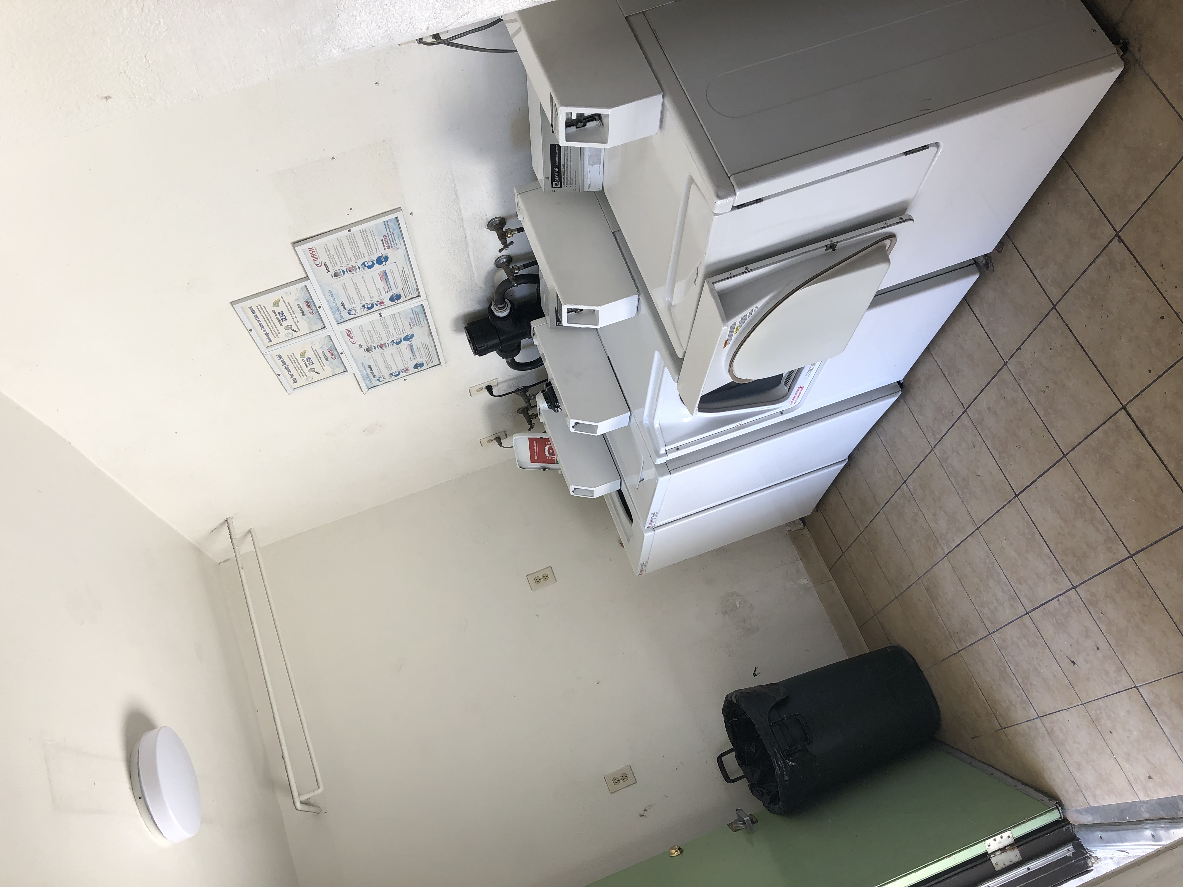Right side view of a laundry, two white washers, two white dryers, beige flooring, big trash can with plastic bag, instruction signs on the wall.