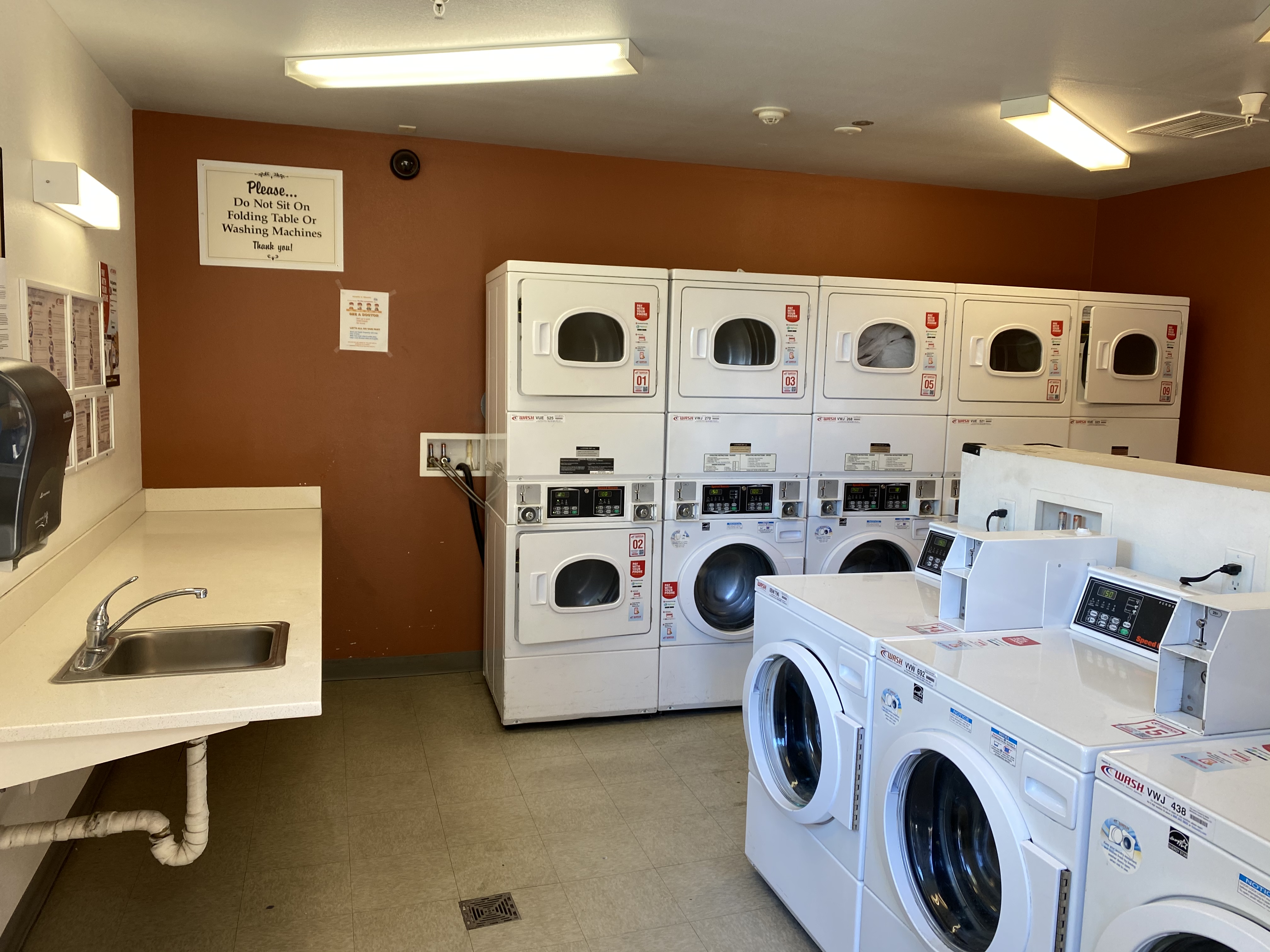 Riverwalk apartments laundry room. ten stacked dryers along wall and washers side by side near center of room. long folding table with sink. security camera in teh laundry room