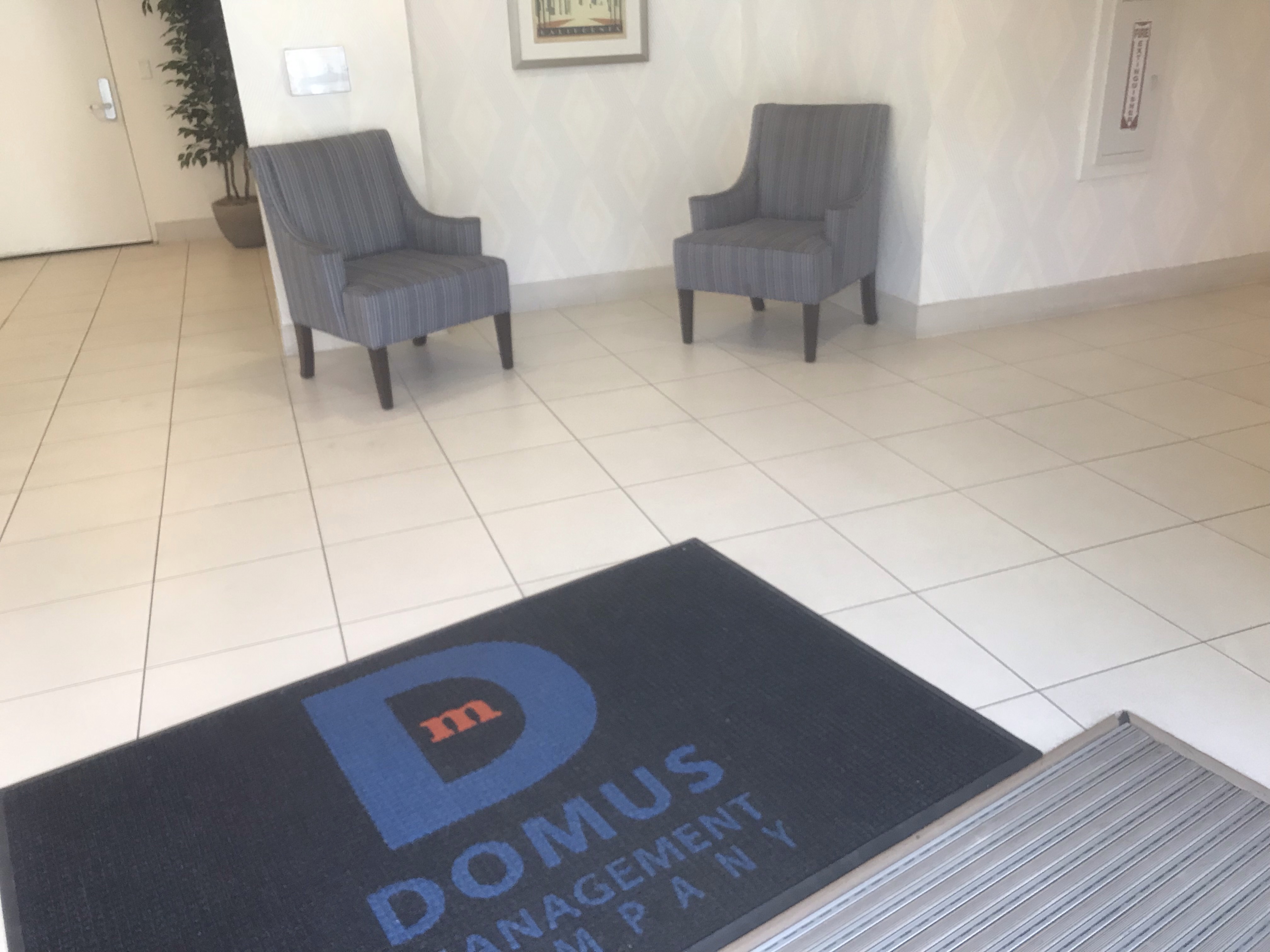 View of a common area, beige tile, two area rugs, one rug with Domus management name on it, two gray upholstered arm chairs, a door, a plant near the door, a fire extinguisher.