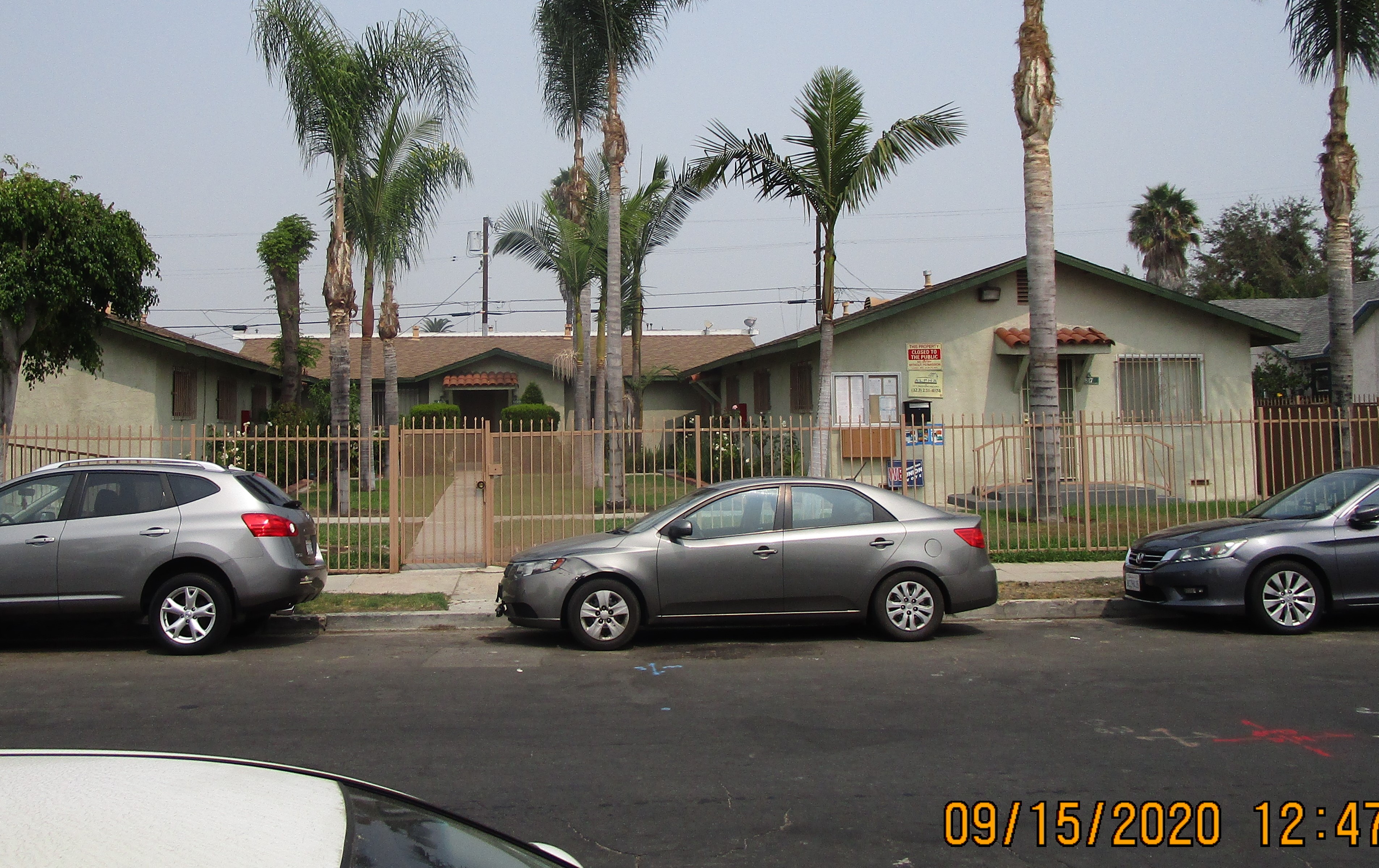 Front view of a light green courtyard style building, brick roof on top of each unit entrance, gated, locked showcase and closed to the public and management signs are posted on the right side of the building, palm trees, bushes and flowers, parked cars i