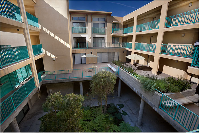 Different view of a courtyard inside a four story building with many plants and trees. The second floor has a lounging area with benches, umbrellas and more plants. The railings on the passageways on each floors are teal.