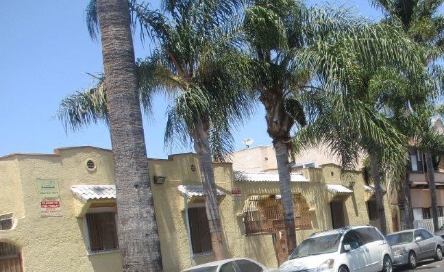 Side side view of a yellow courtyard style building, brown steel gate, multiple gated windows, white brick roof style on top of each window, parked cars, four palm trees in front of the building.