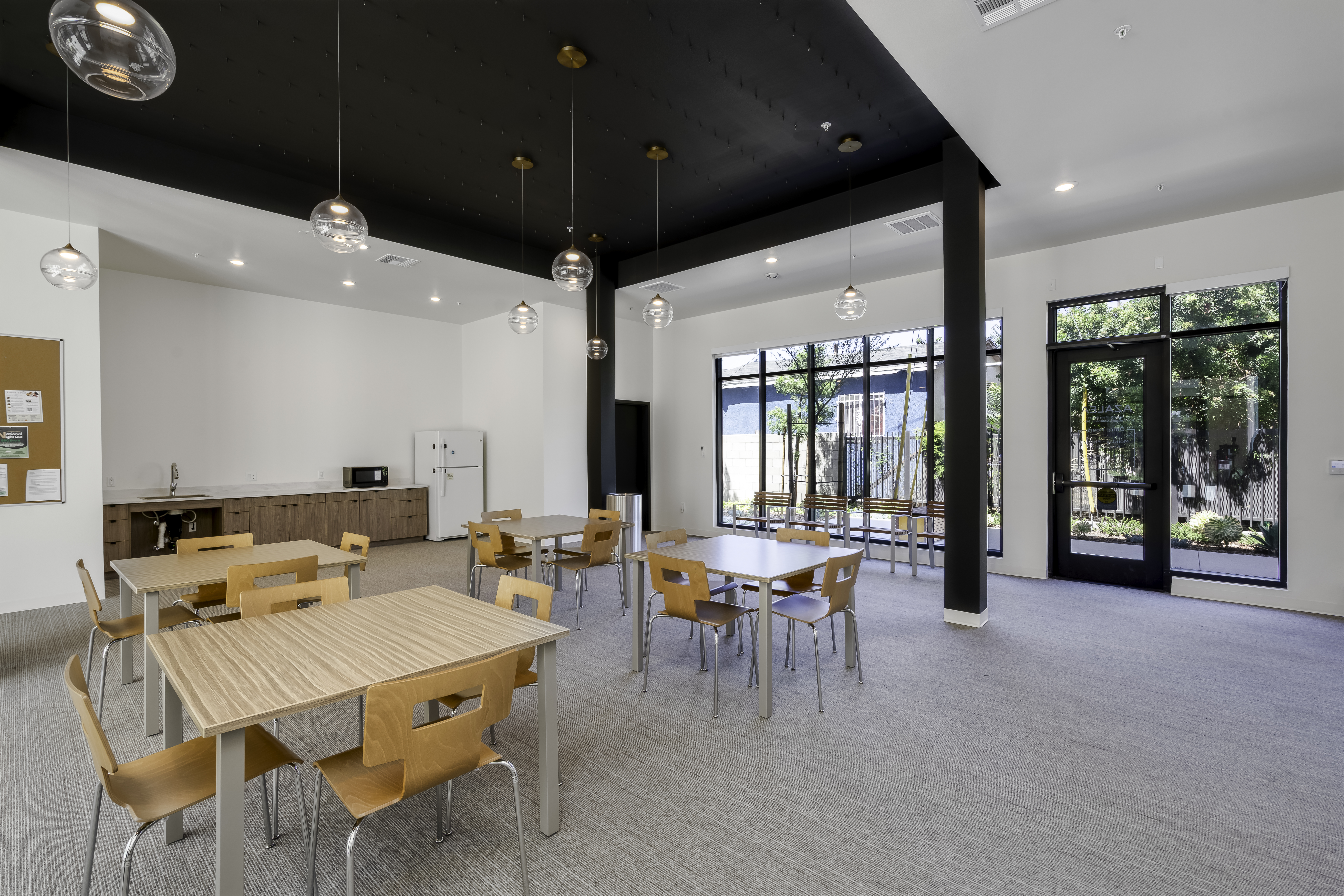 Community Room. With Refrigerator and sink. Four square tables with chairs. A black section of the ceiling with hanging light fixtures with round glass bulb coverings in front of a glass wall showing the outside.