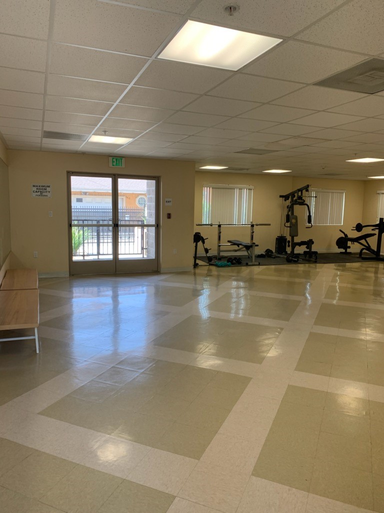 Different angle of a fitness room. There is a bench, a chest press machine, two flat benches for weights. Room is spacious and floor is tiled.