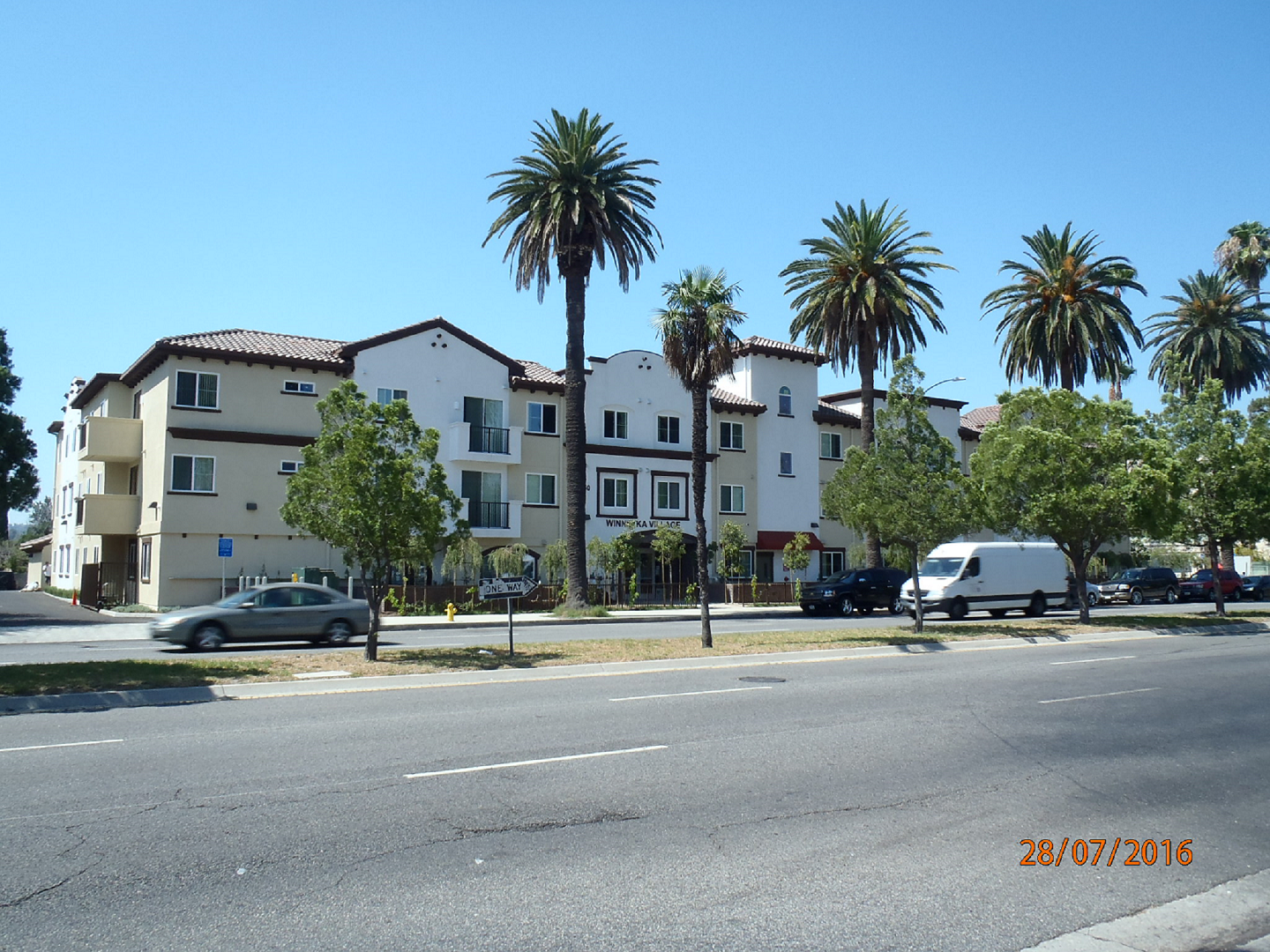 Street view of 3 floor apartment building with Palm trees and trees shrubs on thr sidewalk and cars in the front