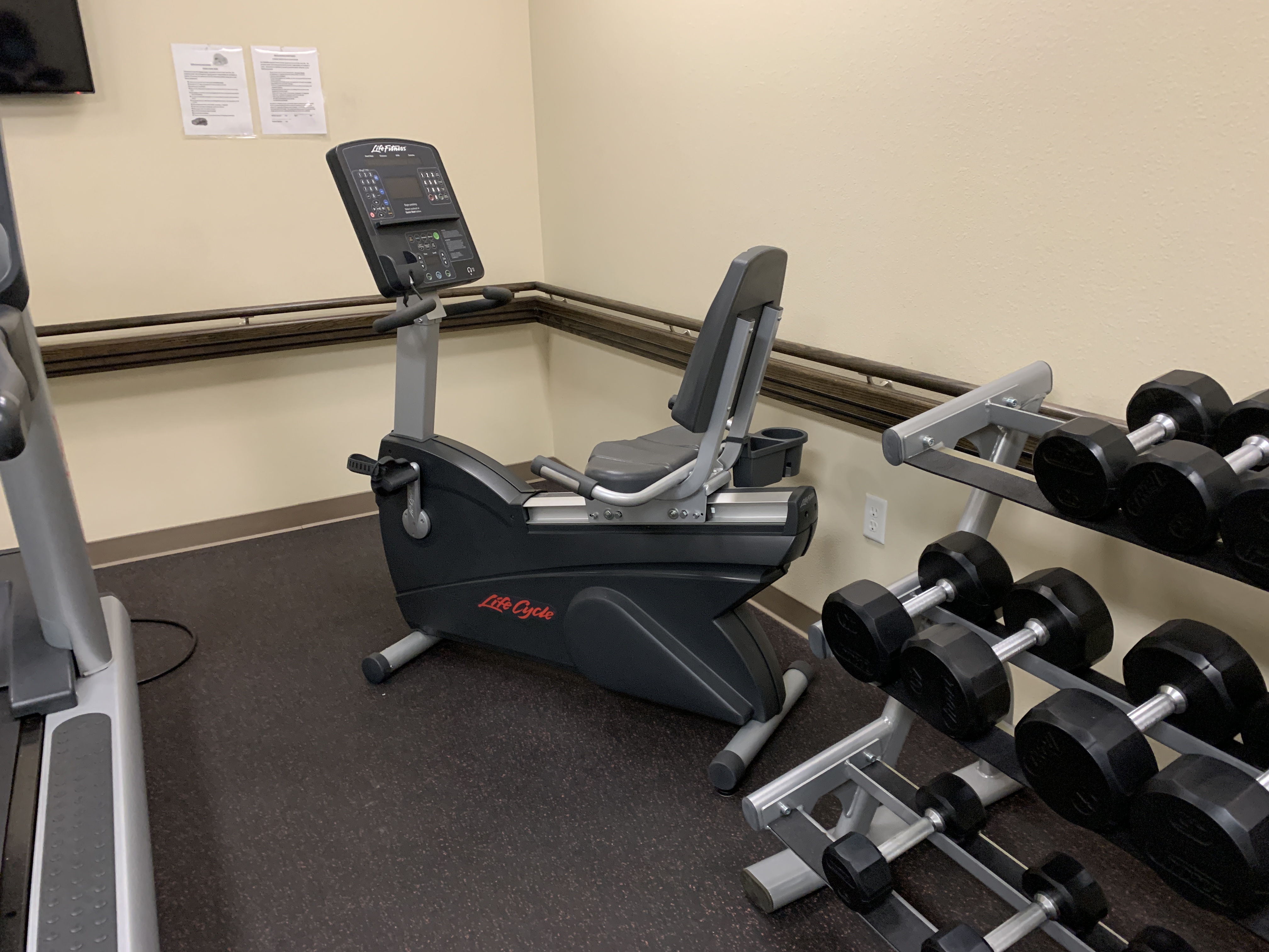 View of a corner of a fitness room. There is an exercise bike machine and weights on a stand.