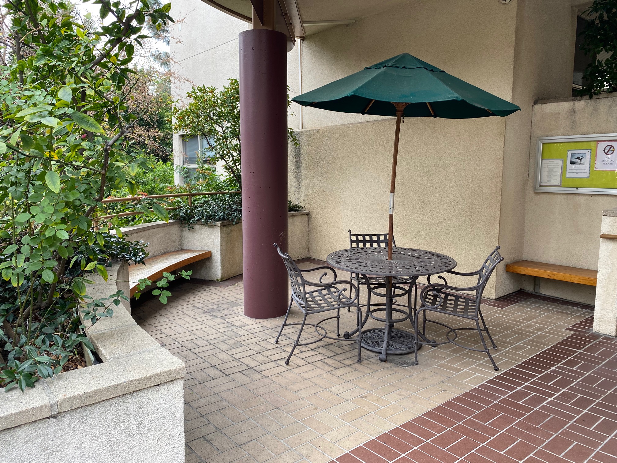 image of la brea franklin seating area. large curved planters with bench seating. in center is patio table and chairs with umbrella. Bulletin board on wall