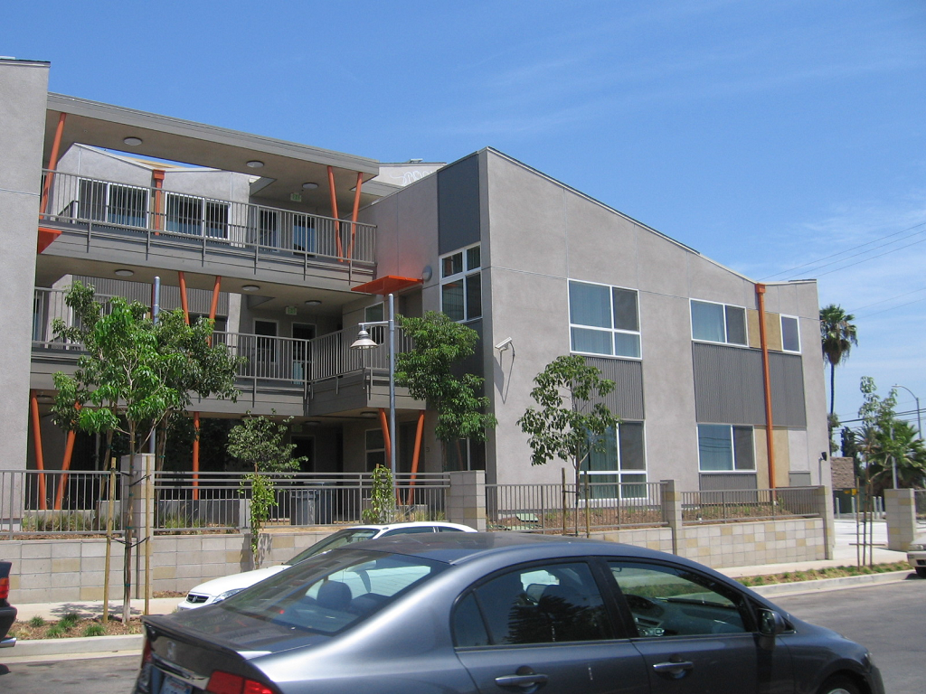 Image of gated gray 3 floor senior apartment building with orange trim and street parking alond building