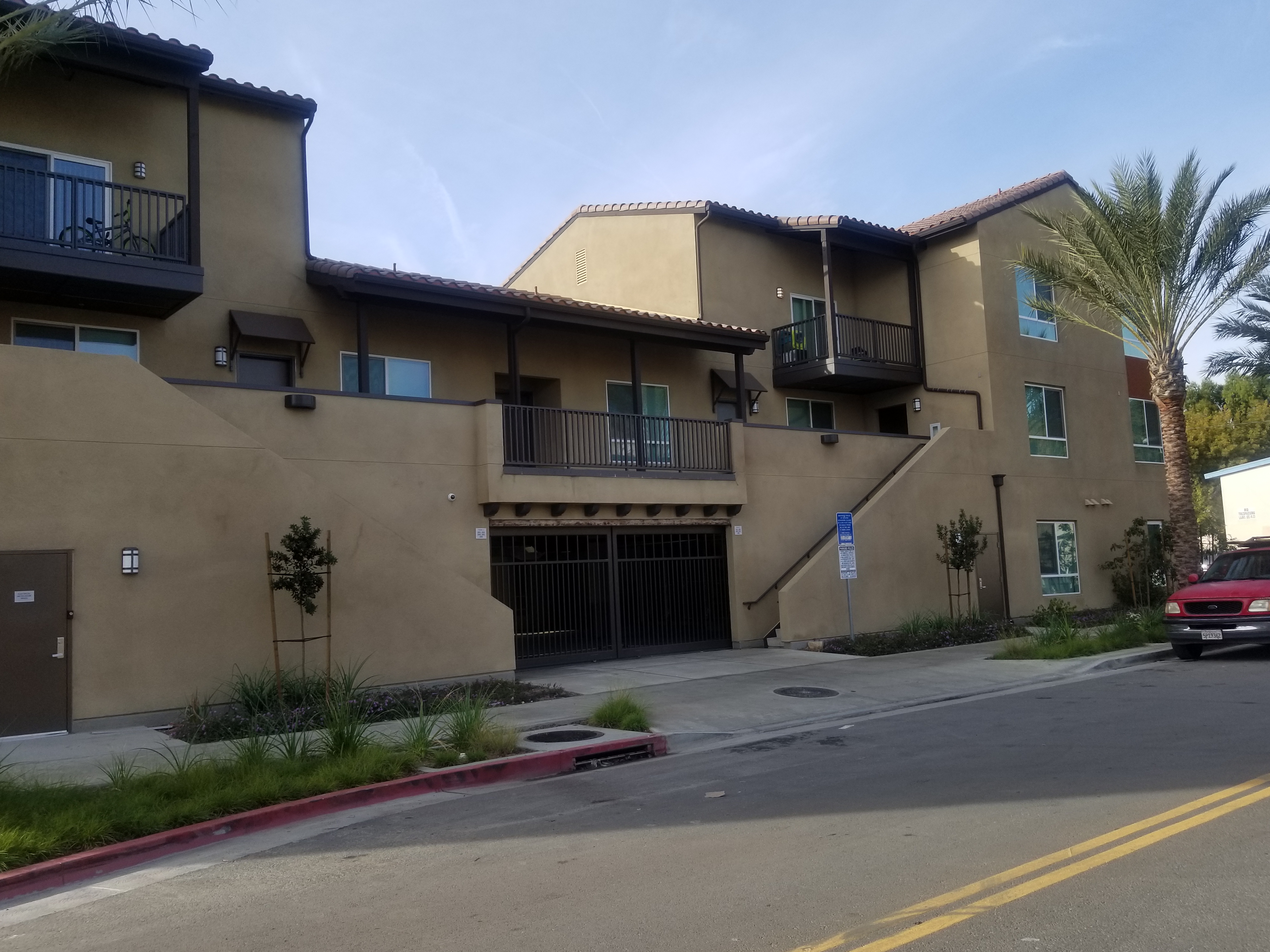 Different view of the outside of a three story light brown building. Ground floor has a gated parking lot entrance. There are newly planted trees, plants and a palm tree in front of the building. There are two staircases across from one another that lead to the sidewalk.