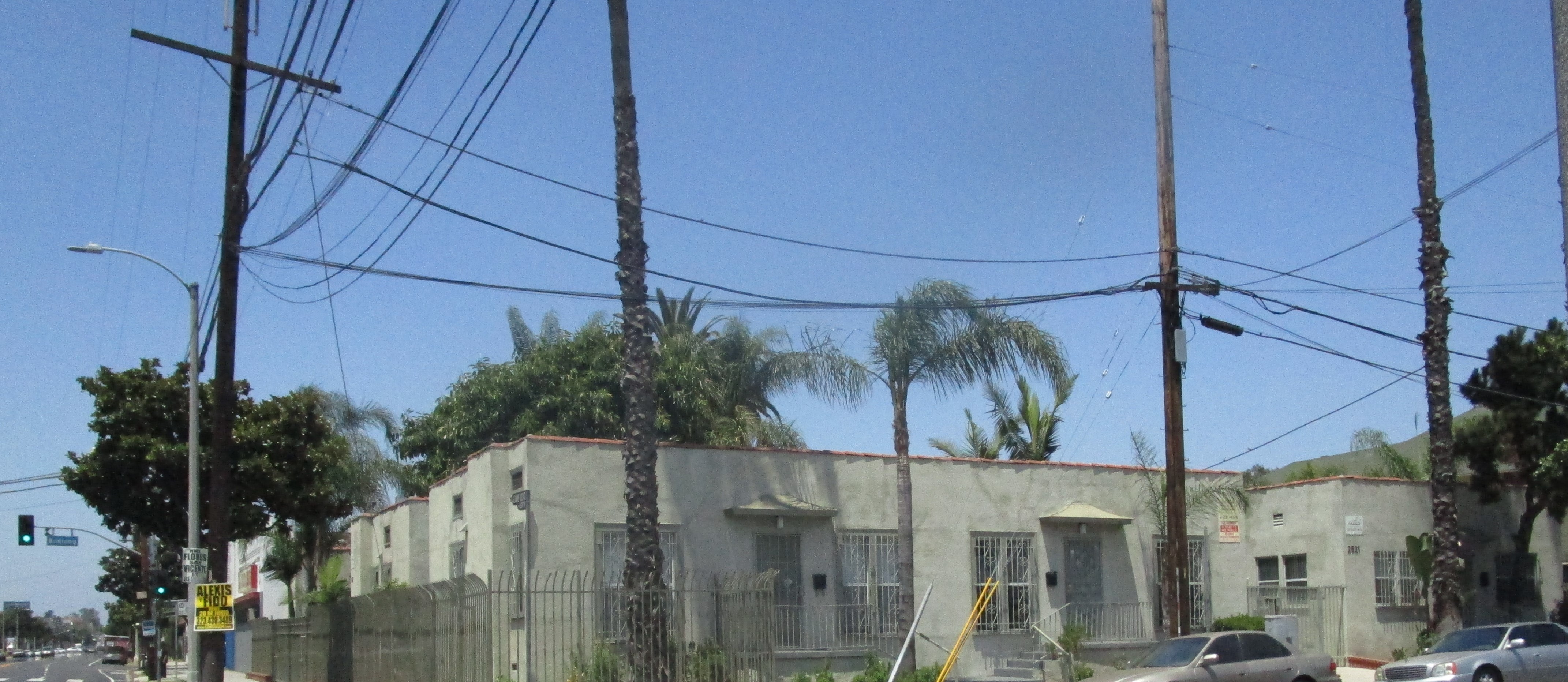 Image of property from corner street view. 
Buildings are light green with fences and a gate. 
Outside facing windows have bars. There are 2 electricity poles and 3 palm trees.