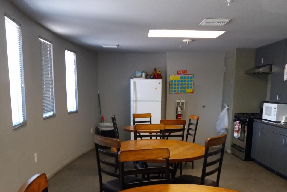 Community Room with three round tables with chairs. A refrigerator, stove, microwave, and kitchen cabinets.