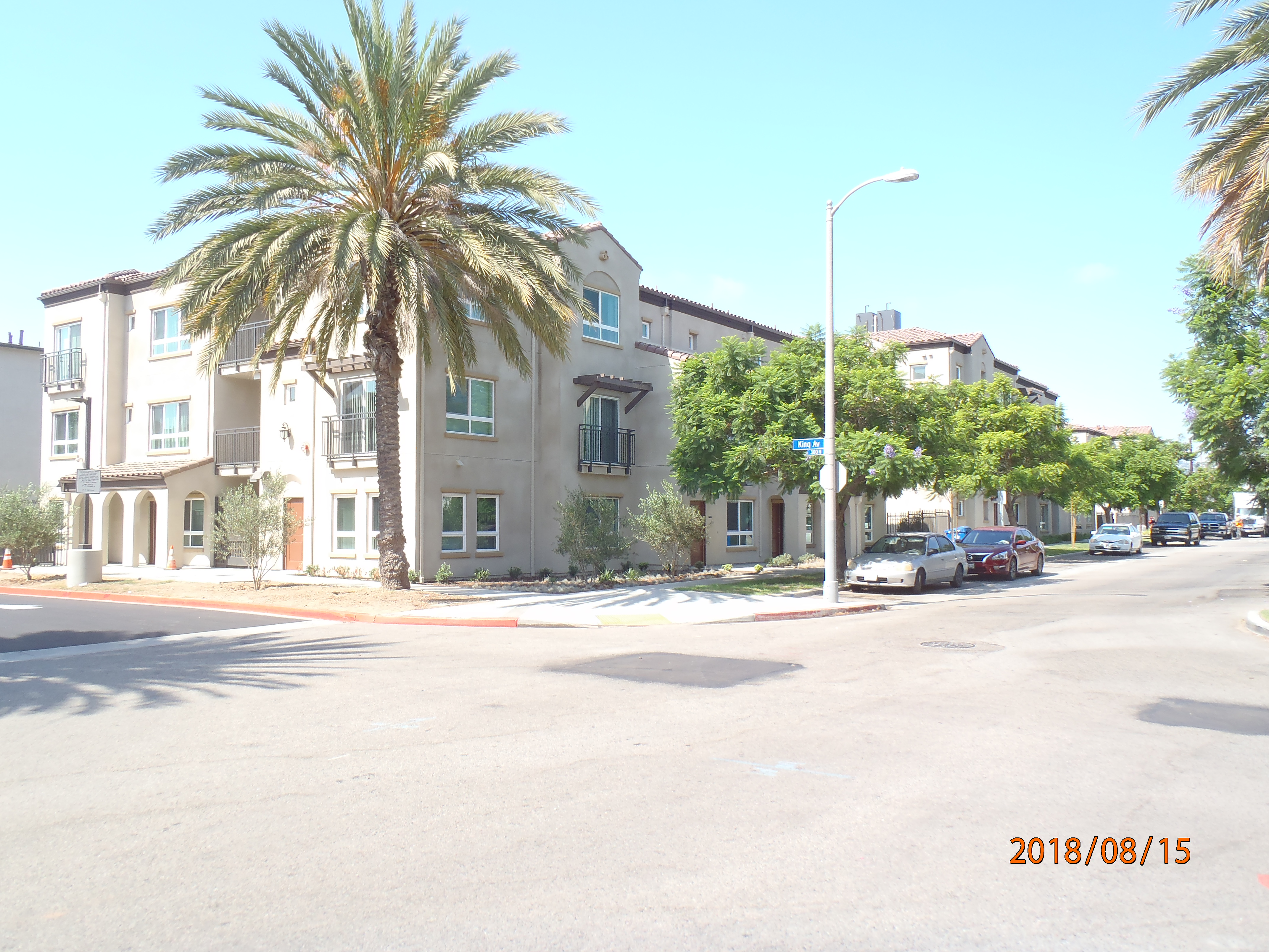 Corner view of a beige and brown three story building, a tall palm tree on the corner, other trees on the right side of the building, multiple windows, some with a brick shade and others with an iron balconies.