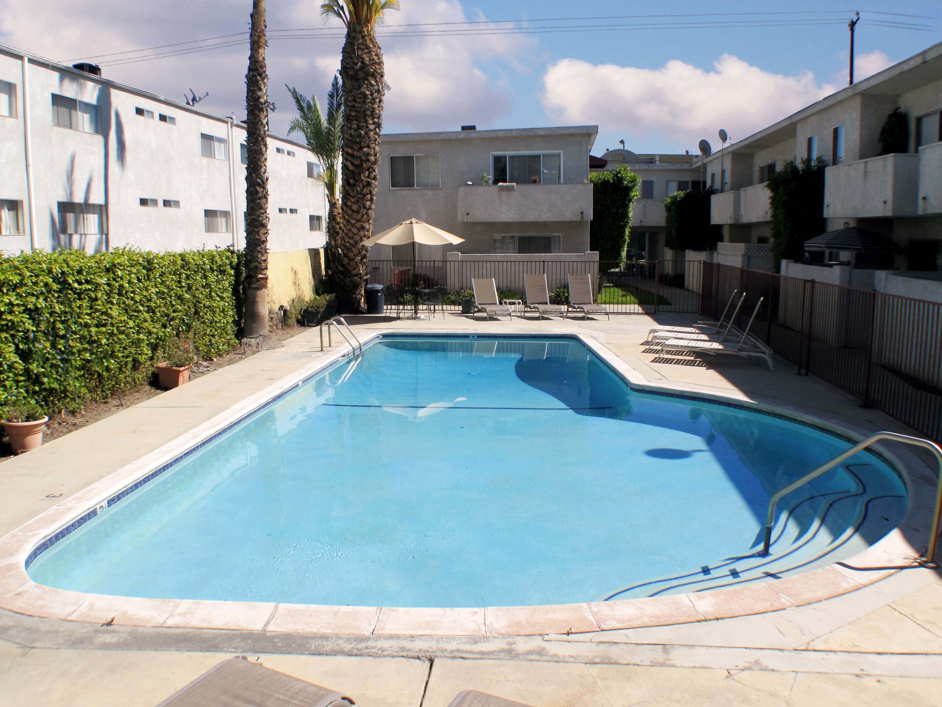 Outdoor pool with lounging chairs, a table with an umbrella, and a hedge on the side. Pool area is fenced. Pool is inside a two story white building.