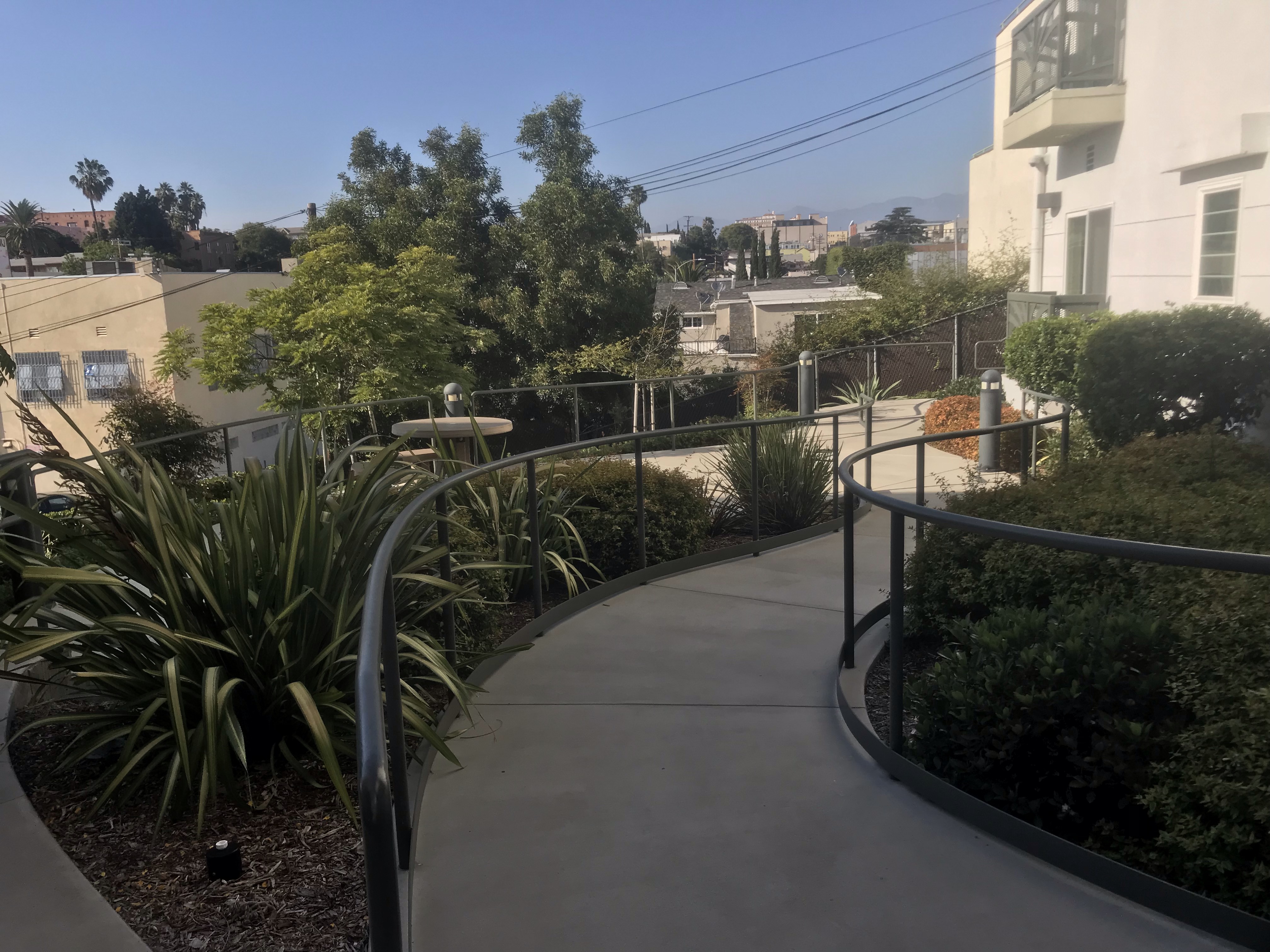 View of a concrete walkway with handrails, a lot pf bushes and plants on both sides.