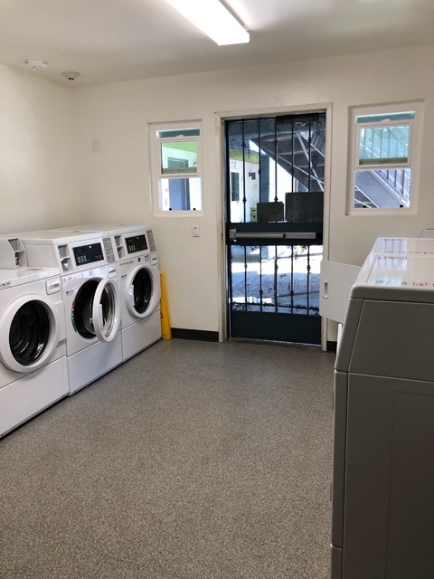 Inside view of a laundry room with multiple washers and dryers across from each other. Entrance is a metal door with a push bar on it. The room also has two small windows.