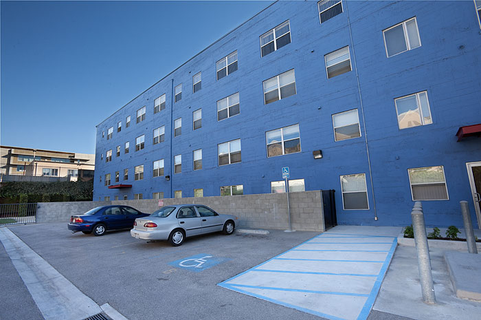 View of an outdoor parking lot for a blue four story buikding.There is a gate for entry, and an accessible space near the building's entrance.