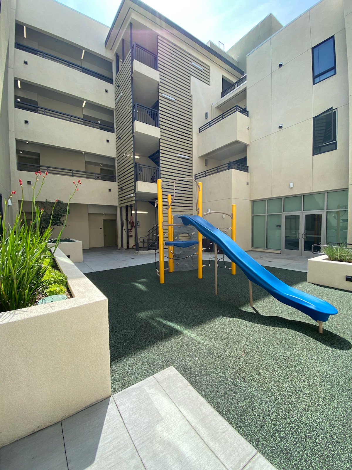 playground in the center of green floor. Planter to the left, staircase for building at the far end.