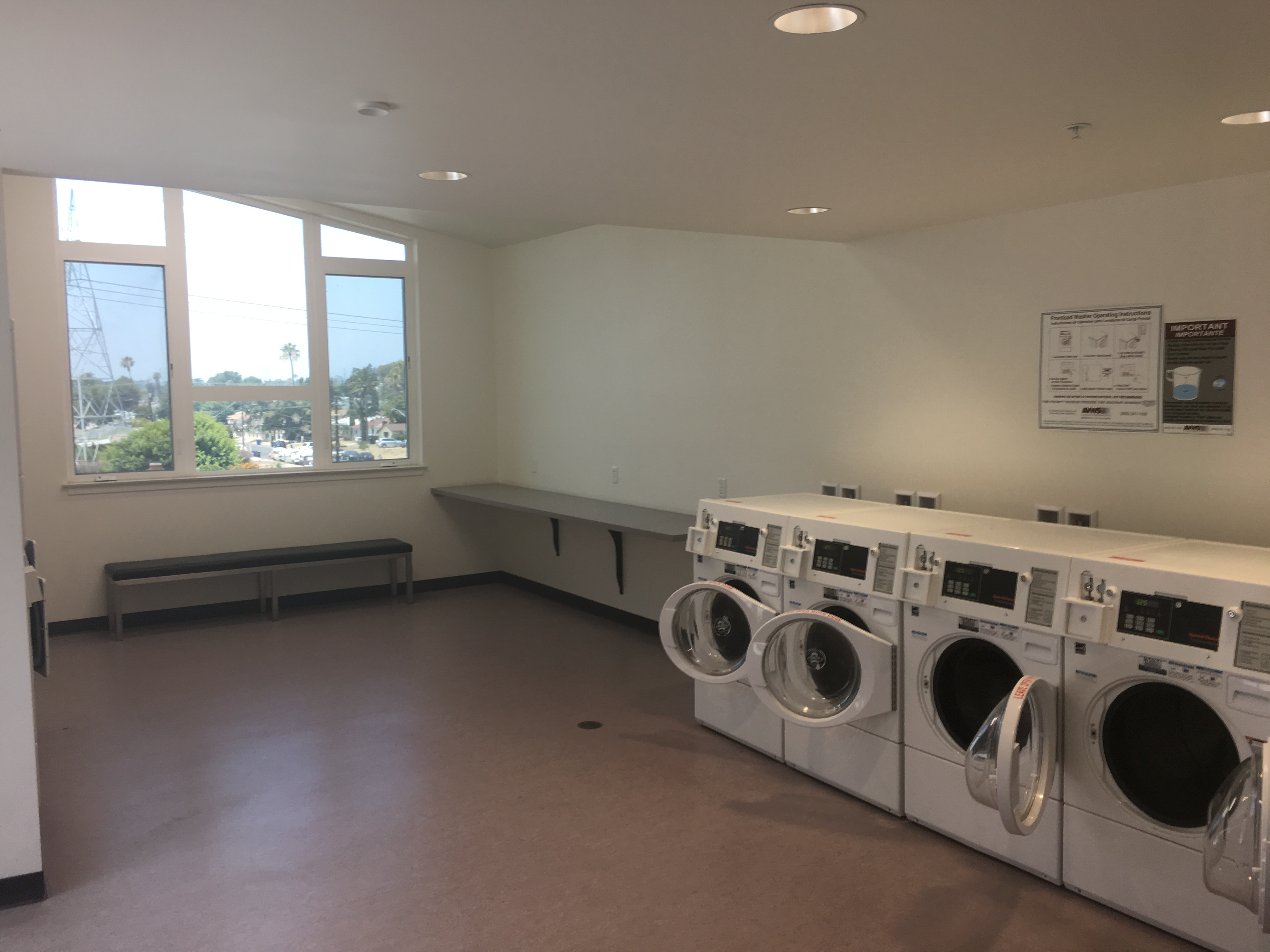interior view of the fourth floor Laundry Room in the PSH Campus showing four washers against the wall next to a long table attached to the wall. To the side of the table is a long bench to sit with a large window above sectioned in six panels