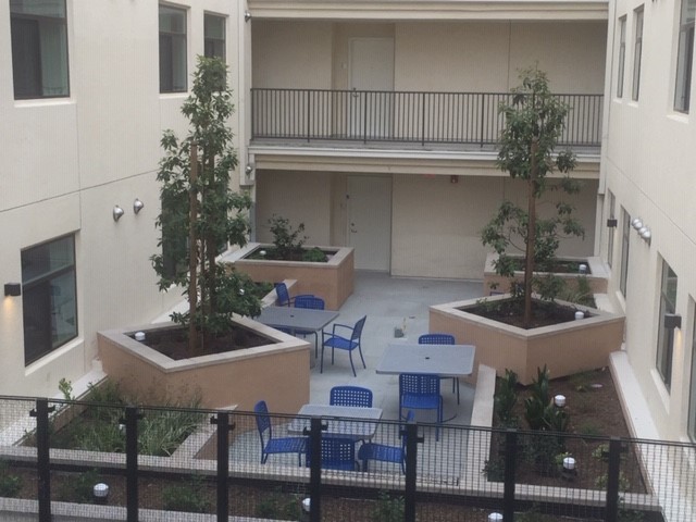 Exterior view of the Pico Robertson Senior Community Apartments courtyard in the middle of the building. 3 grey tables with blue chairs. 4 large cemented planters, each with a tree