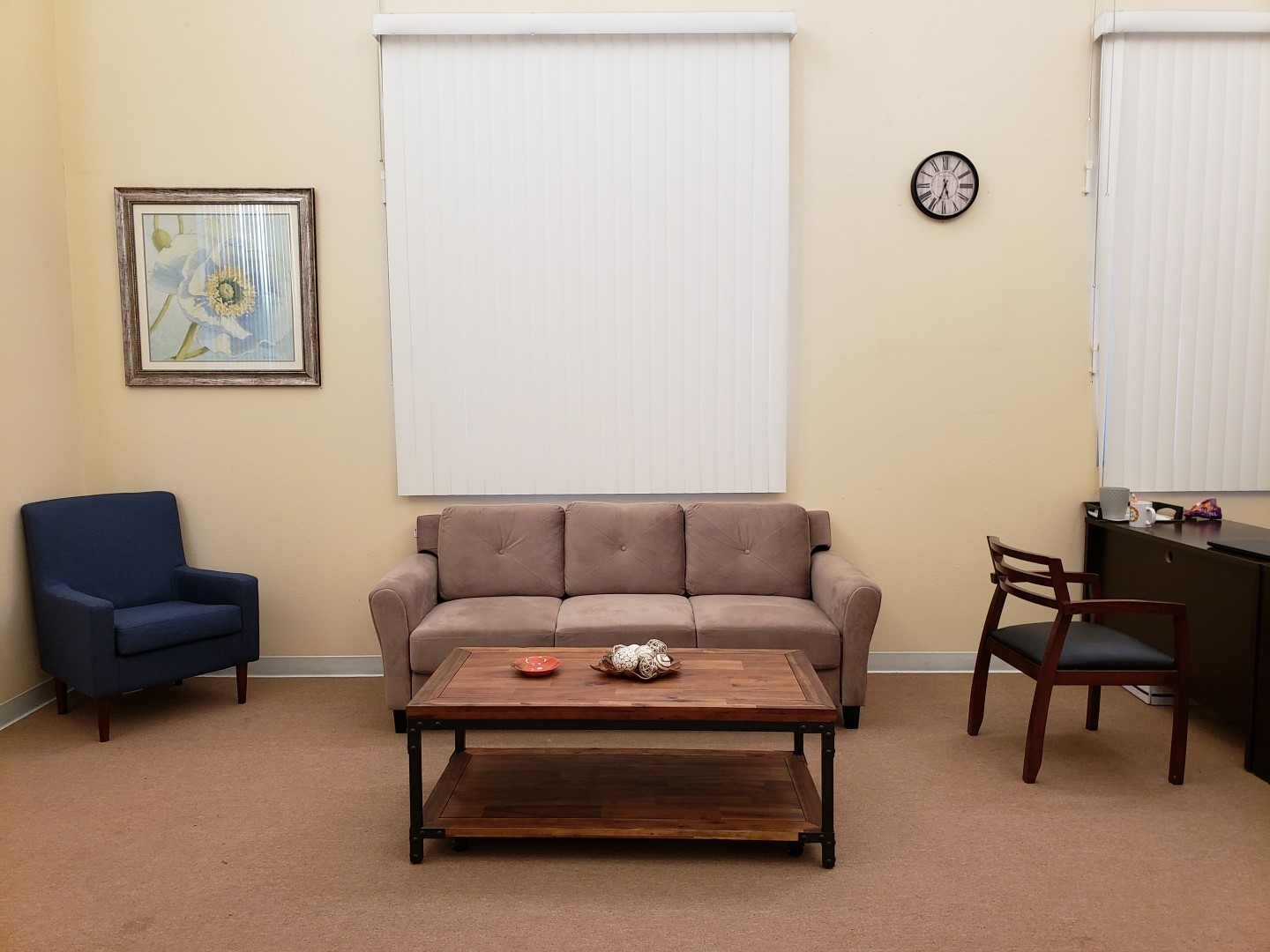 Lobby area with a sofa chair, couch, coffee table, and reception desk. There are two windows with vertical blinds. On the wall there is a clock and a painting, and the floor is carpeted.